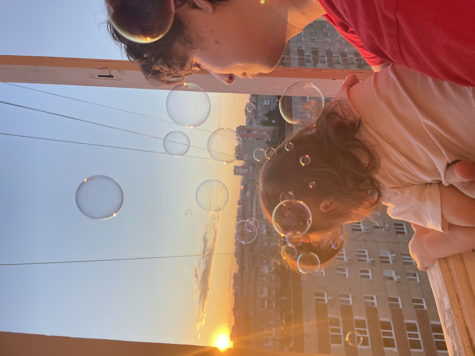 Two individuals, a young adult and a child, are observed from a high vantage point, likely a balcony or window, overlooking the city of Izhevsk, Russia, at sunset. The setting sun, a bright orange orb, is visible on the far left horizon, casting a warm, golden light across the entire scene and the urban landscape.
The young adult, positioned on the right, is seen in profile, facing left, with dark, curly hair and wearing a red garment. Their skin is illuminated by the intense sunset light. In front of and slightly below them, a child with dark hair tied back, wearing a light-colored top, also faces left, looking out over the city. The child's back is mostly to the viewer, and one arm rests on a light-colored wooden railing. The larger individual appears to be supporting or holding the child.
Numerous iridescent soap bubbles, varying in size, float in the air around and between the individuals, reflecting the vibrant sunset colors. Some bubbles have distinct glints of orange light. Beyond the balcony, a dense urban environment stretches into the distance, characterized by multi-story residential buildings with multiple windows. The sky transitions from a fiery orange near the horizon to a clear light blue higher up, with a few faint clouds visible. Thin, vertical lines, possibly clotheslines or structural wires, extend upwards from the balcony area into the sky.