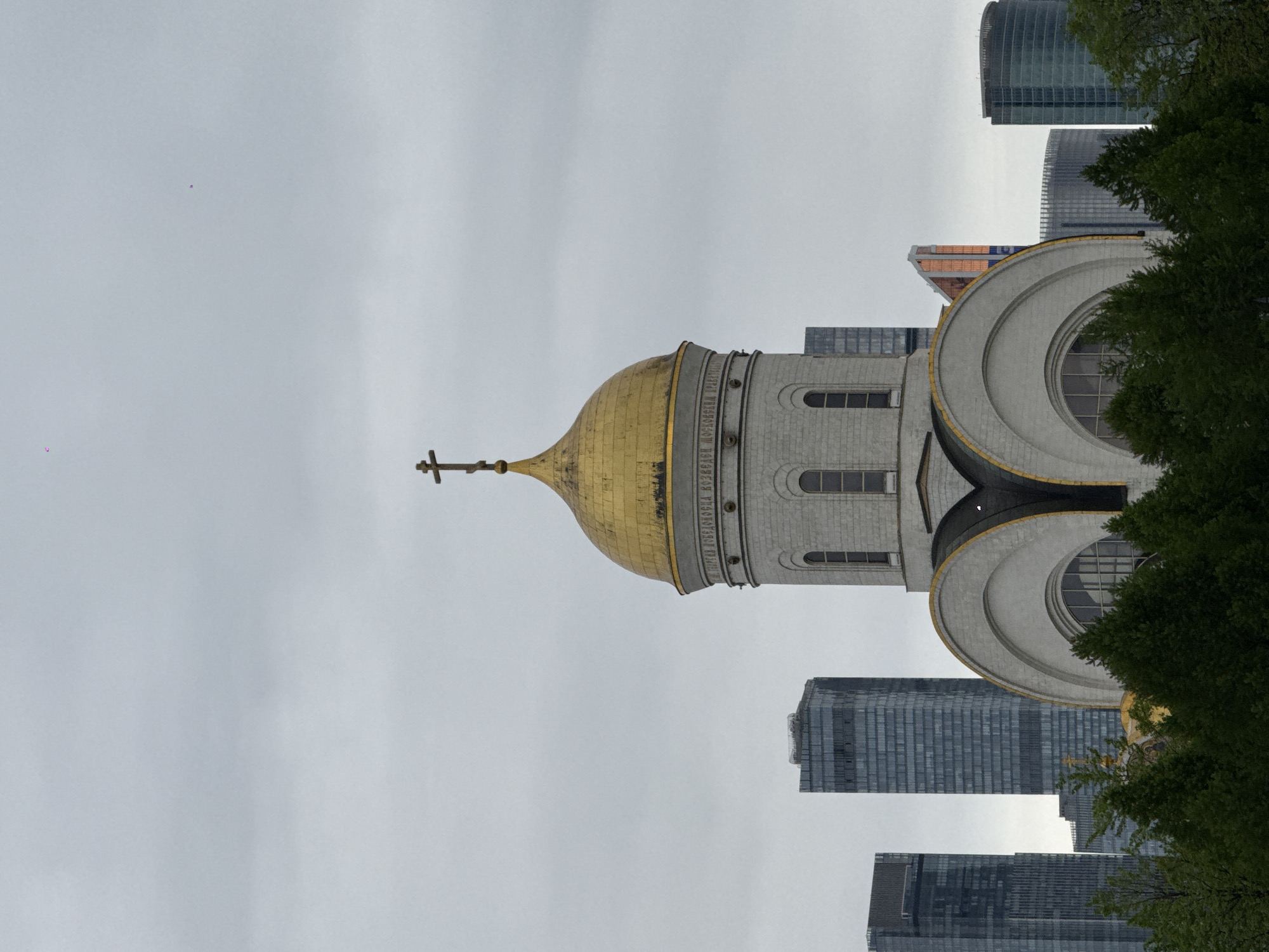 The image depicts the Cathedral of Christ the Savior in Moscow, Russia. The cathedral features a large golden dome with a prominent cross. Its architecture includes arched windows and decorative elements. In the background, modern skyscrapers rise above the cathedral. Parts of trees are visible, suggesting the cathedral is viewed from a park. The sky is overcast.