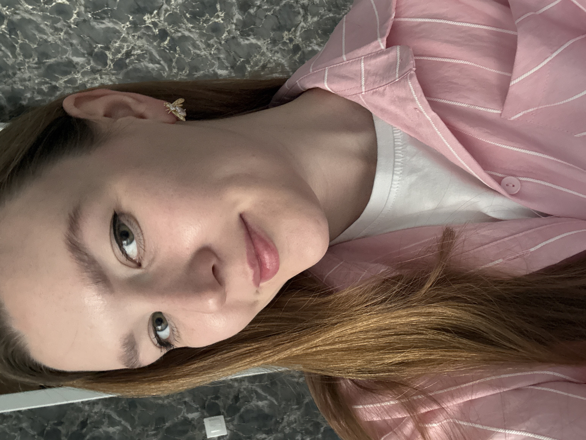 A young woman with light skin, light eyes, and auburn hair is posing for a selfie. She is wearing a white t-shirt under a pink and white striped shirt. In her left ear, she has a gold-colored earring shaped like a bee. The background is a dark, mottled surface, possibly a counter or floor. The lighting is diffused and even. The location in Izhevsk, Russia is unknown.