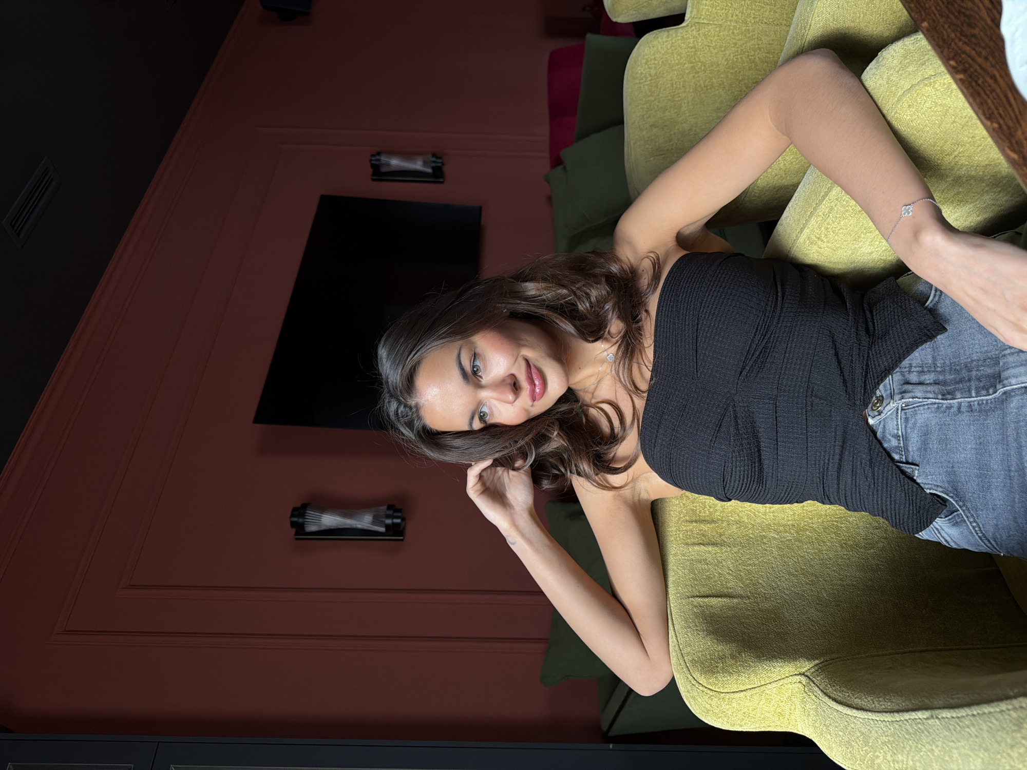 A woman is reclining in a green armchair indoors. She wears a black top and jeans. The wall is a shade of red and has two lights and a television mounted to it. A glimpse of additional seating, including pink chairs, can be seen in the background. The location is likely Krasnoyarsk, Russia.