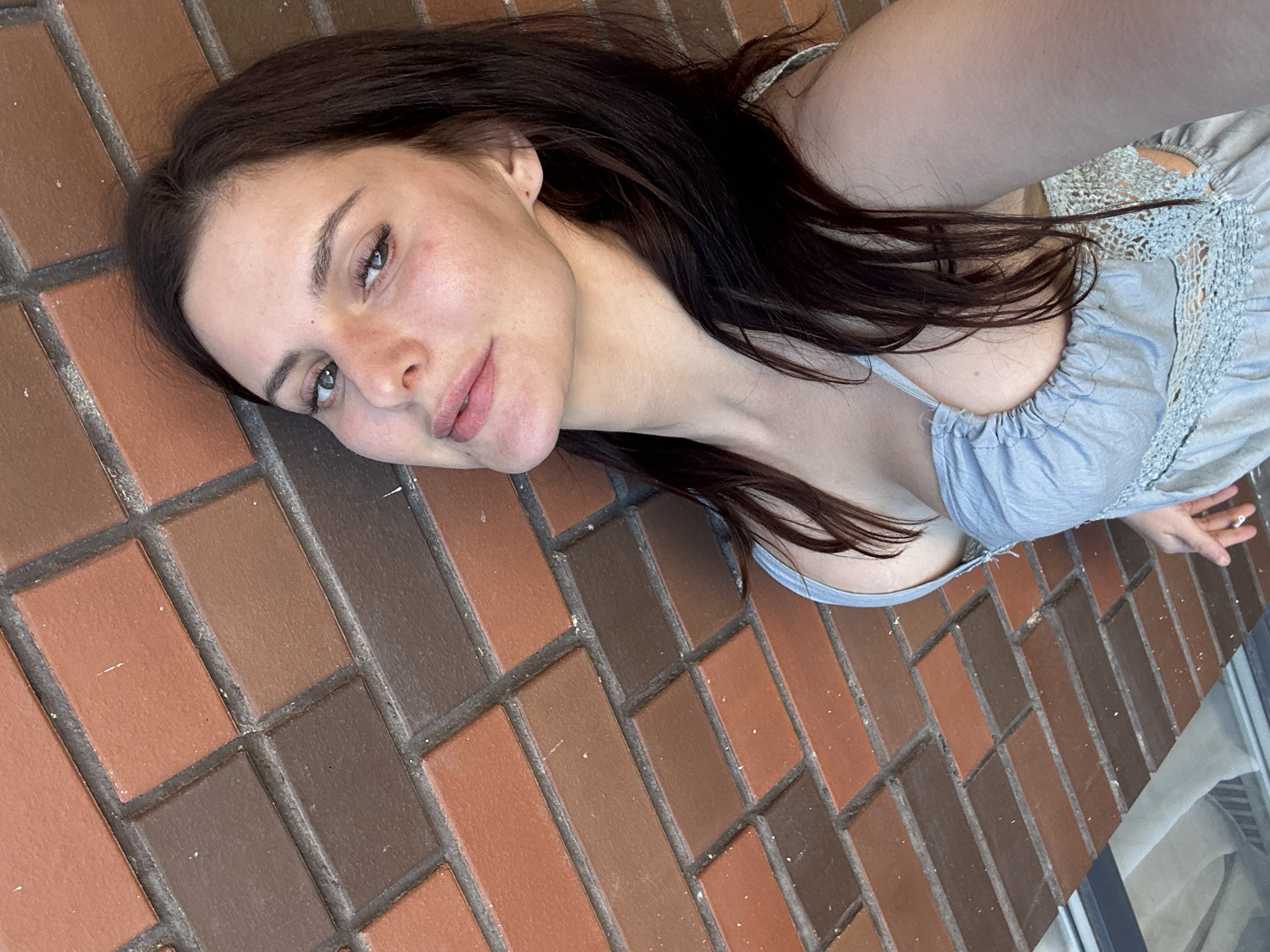 A woman with dark hair and fair skin poses for a selfie against a brick wall. She is wearing a light blue sundress with lace trim. The wall is made of bricks of varying shades of red and brown. The lighting suggests it is daytime. The image is cropped to focus on the woman's face and upper body, with the brick wall occupying the background.
