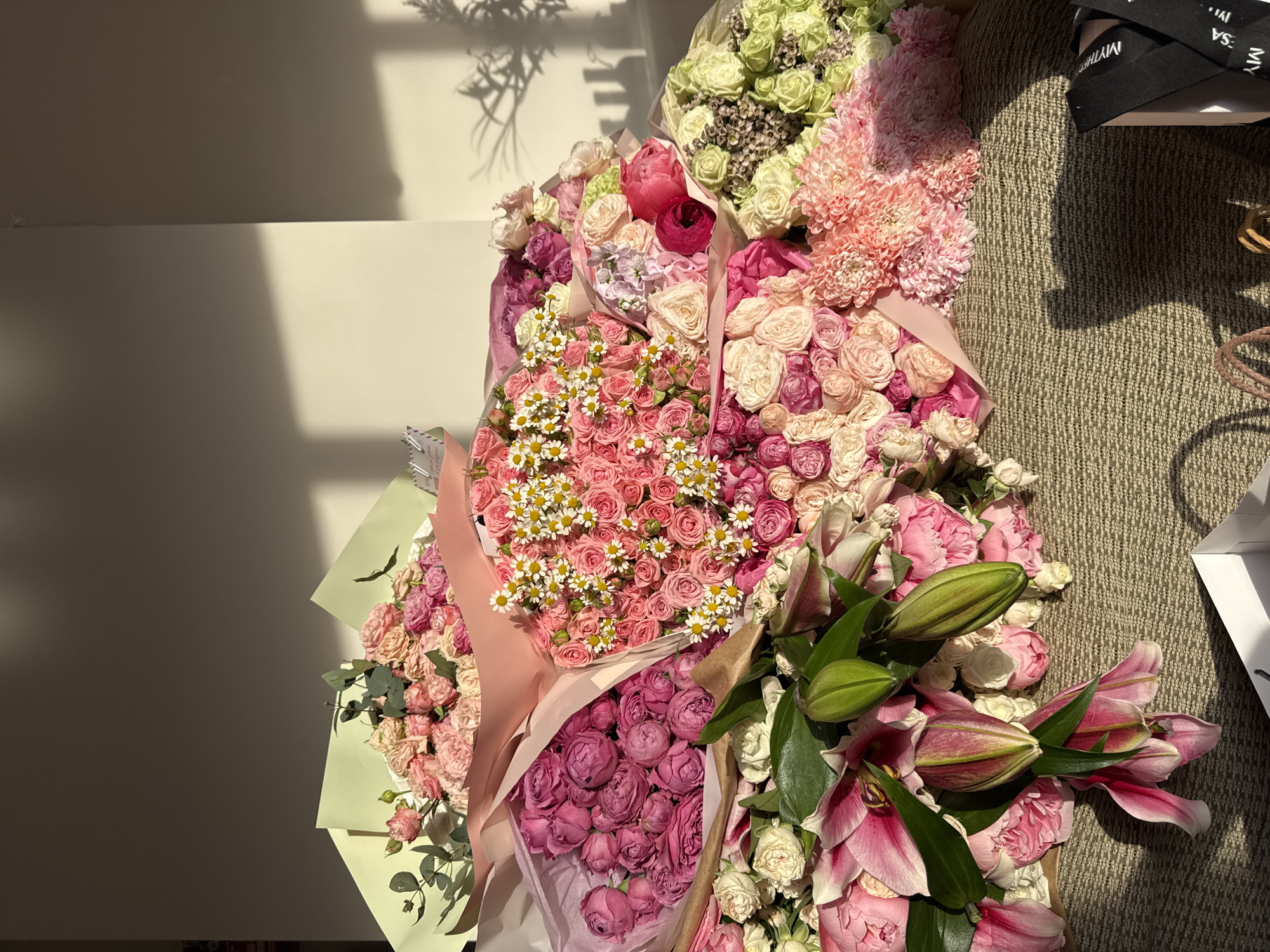 The image shows an overhead view of several bouquets of flowers on a textured surface, potentially a rug. The bouquets consist of various types of flowers, including roses, lilies, and daisies, in a range of colors, primarily pink, white, and green. Some of the bouquets are wrapped in paper of different colors. A pair of black sandals with white lettering are visible in the upper right corner, near a white box. The setting appears to be indoors, with natural light coming in from the left side of the frame, casting shadows on the wall in the background. The city and location are not identifiable from the image.