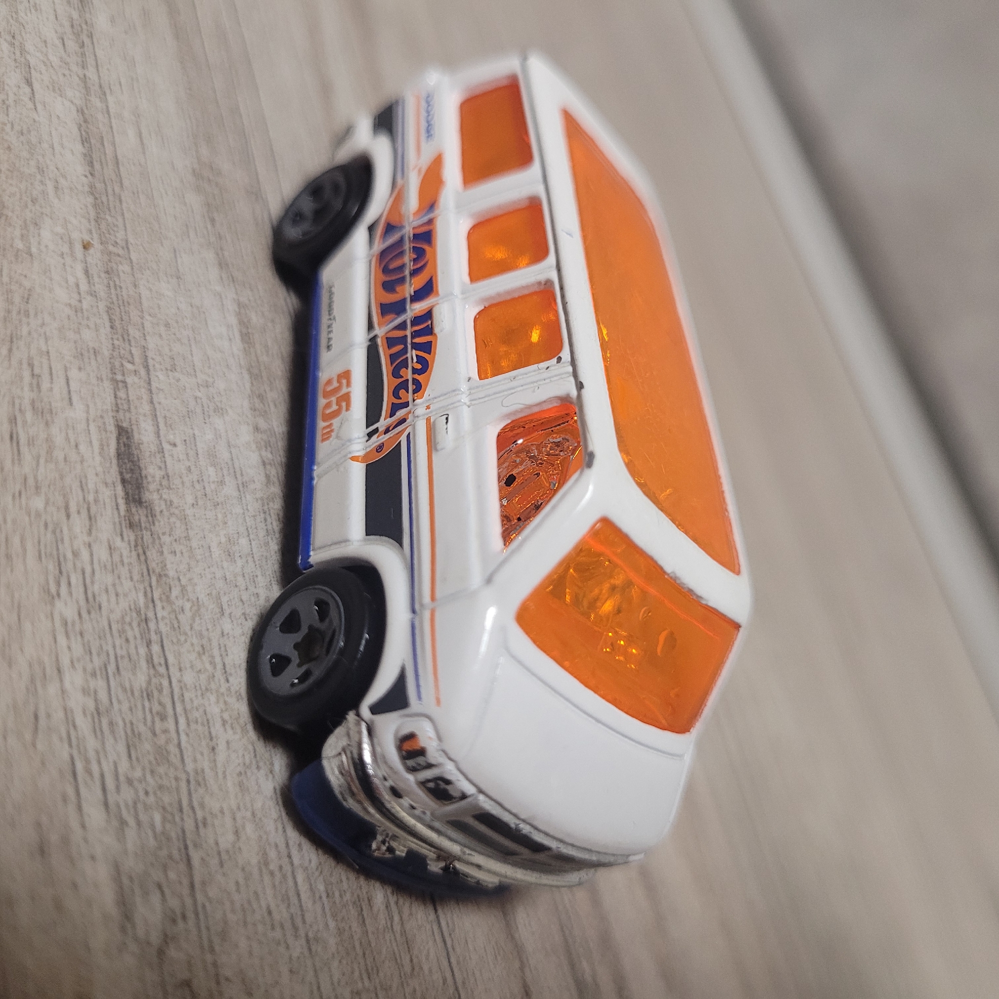 A white toy van with orange tinted windows and roof is shown from a slightly elevated angle, resting on a light-colored wooden surface. The van has the "Hot Wheels" logo and "Dodge" branding on its side, along with racing stripes and the number "55th." The front of the van shows a chrome bumper and headlights, and the wheels are black. The scene is indoors, likely on a table or floor. There are no people or any other objects visible. The lighting appears to be artificial, casting soft shadows.