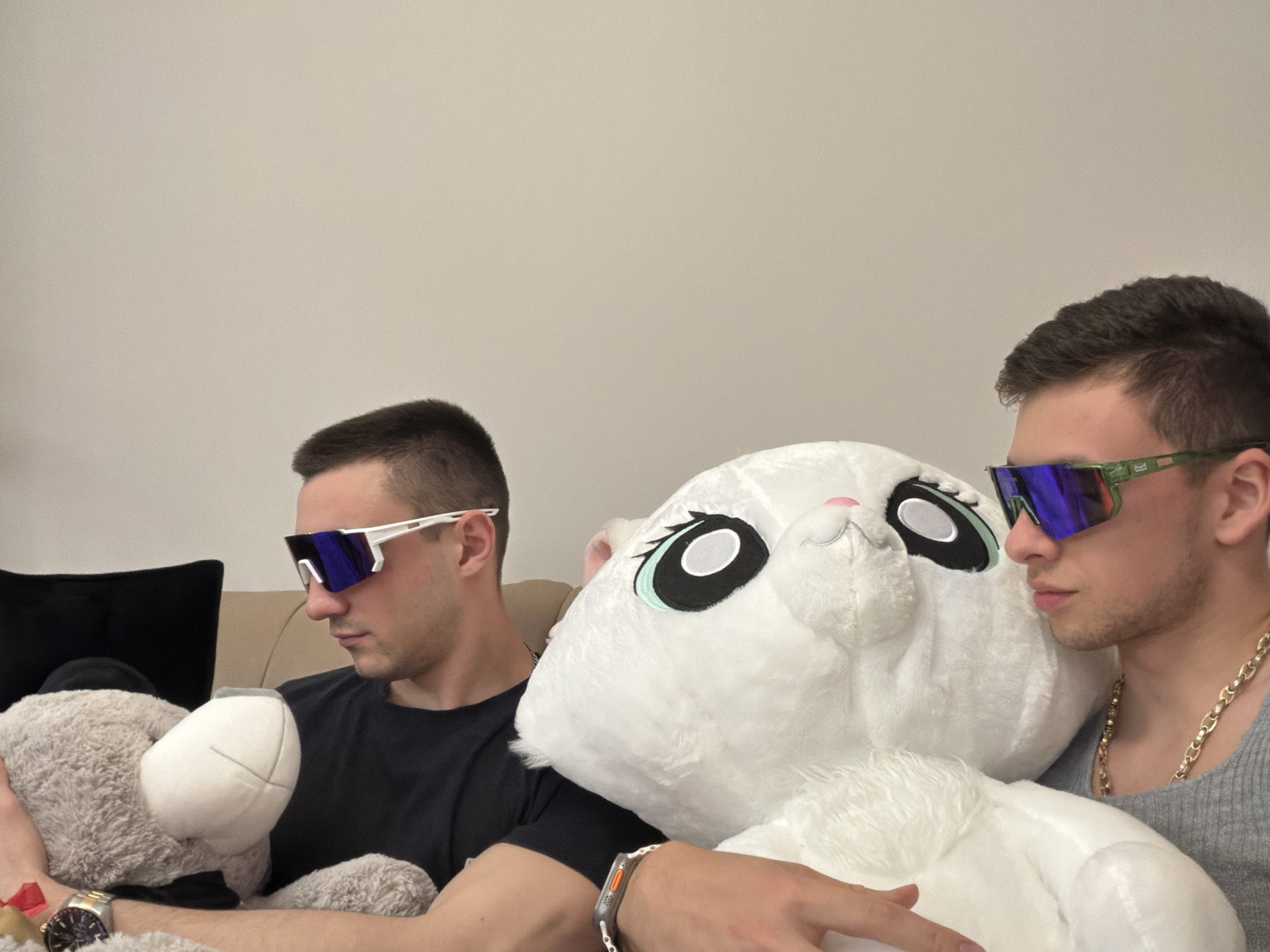 Two young men are seated indoors on a light-colored sofa, each accompanied by a large plush toy. The man on the left has short dark hair and wears a black t-shirt. He is wearing white-framed, mirrored sunglasses with blue lenses and a silver watch on his left wrist. He holds a large grey plush bear, looking downwards towards it. Behind him, a black cushion is visible on the sofa.
The man on the right also has short dark hair and light facial stubble. He wears green-framed, mirrored sunglasses with blue lenses, a grey ribbed tank top, and a prominent gold chain necklace around his neck. An Apple Watch Ultra is visible on his right wrist. He is looking towards the left.
Between the two men, and partially held by the man on the right, is a large white plush rabbit with big black and teal eyes and a pink nose. The background is a plain, light-beige wall, suggesting a simple indoor setting, possibly a living room. Both men appear relaxed, their expressions obscured by their sunglasses. No visible text is present in the image.