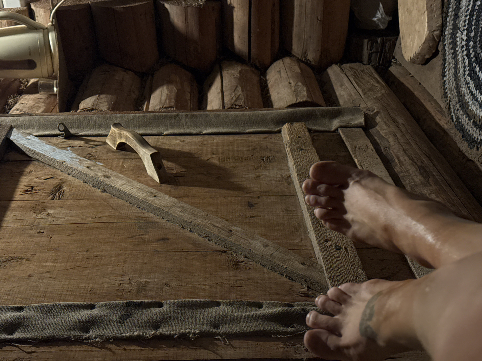 The image shows a person's feet resting on a wooden surface inside a sauna or bathhouse in Nizhnyaya Chermoda, Russia. The wooden structure appears to be a bench or a platform within the room. The logs of the walls are visible. A wooden tool and a container sit on the ledge next to the log wall. A mat sits on the log wall on the right. The feet appear damp or oily. There is a tattoo on one ankle.