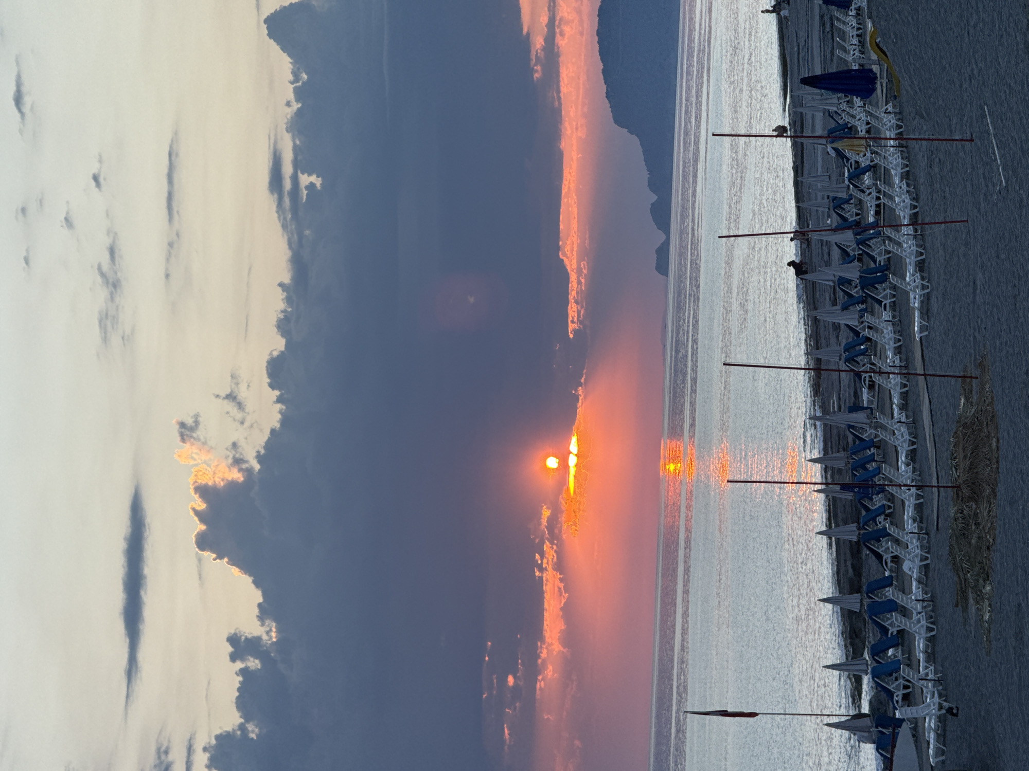 The image depicts a beach scene at sunset. The sun is partially obscured by clouds, creating a vibrant orange and pink sky. The sea reflects the colors of the sunset. A row of beach umbrellas and lounge chairs lines the shore. The location is a beach near mountains with an Unknown City.