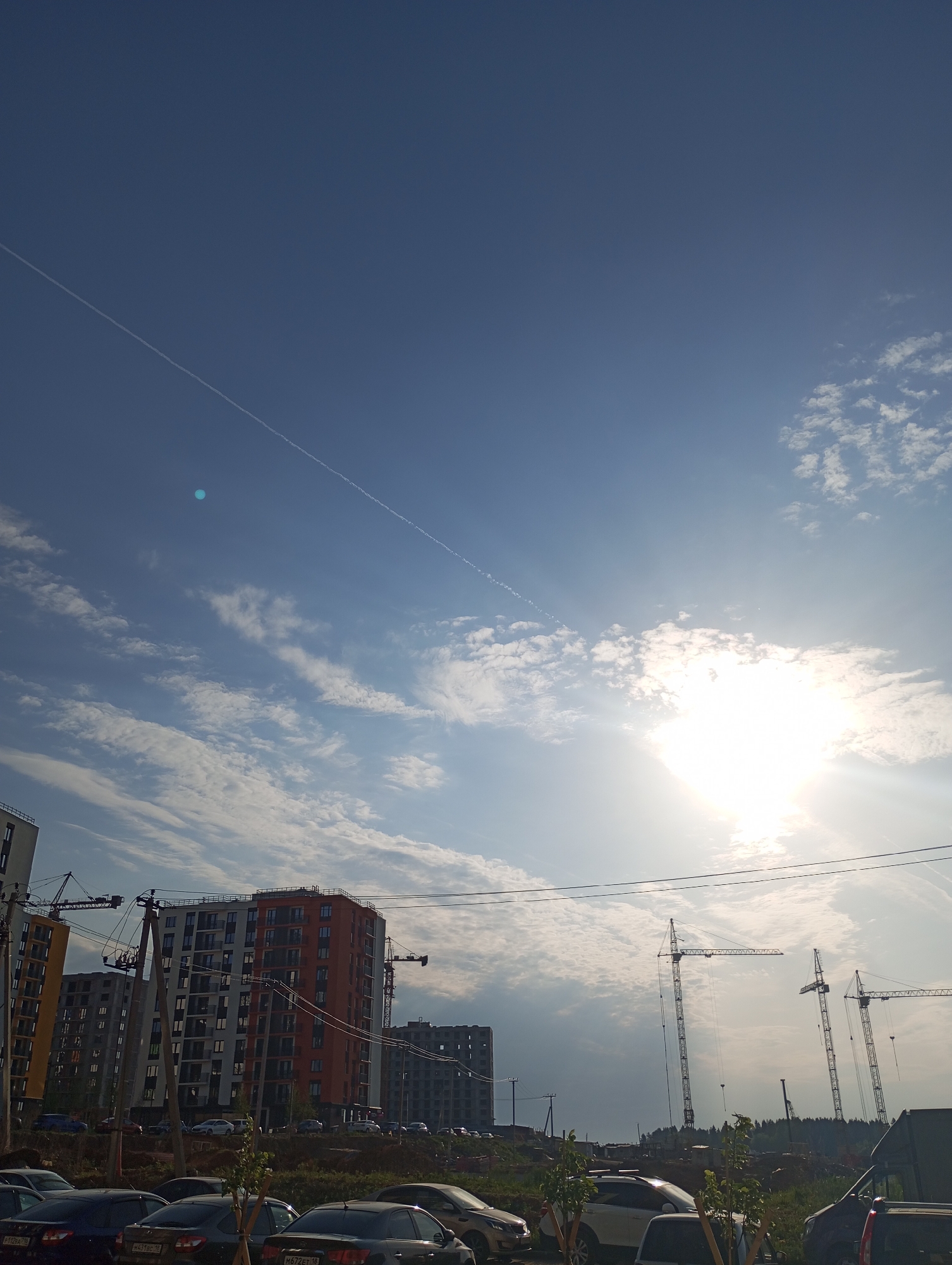 The image shows a daytime scene in what appears to be a construction zone in Pervomaysky, Russia. The sky is predominantly blue with scattered clouds and a bright sun. Two visible contrails cross the upper part of the frame. In the foreground are several parked cars. Behind them is a raised area of land. In the middle ground, several multi-story buildings are under construction, with cranes visible alongside them. Power lines run across the middle of the frame.