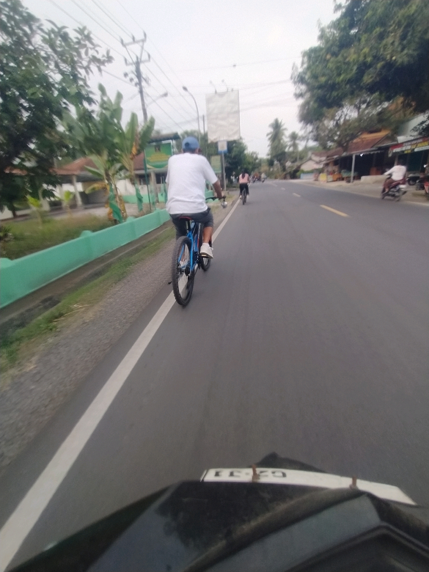 A man wearing a white t-shirt and blue cap is cycling on the right side of a paved road in Desa Karangbenda, Indonesia. He is wearing dark shorts and white shoes. The man is positioned in the middle ground of the image, and his bicycle has a blue frame and black tires. The road is lined with a green fence on the left side, and the surrounding environment includes trees, buildings, and power lines. In the foreground, the front of a dark vehicle is visible, with a white plate displaying black lettering. Other people on bicycles and motorcycles are also present on the road in the background, and some shops and houses line the street on the right side. The sky is overcast.