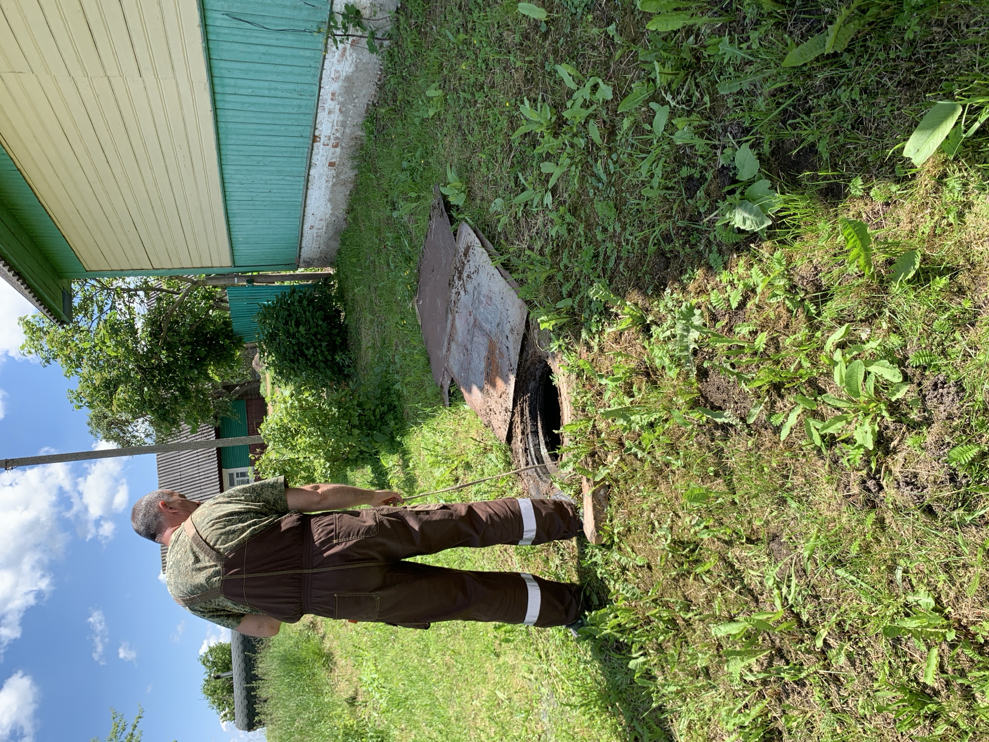 A man wearing brown overalls with reflective stripes on the lower legs stands in a grassy yard near a building with light yellow and green siding. He is holding a long, thin object, possibly a stick or a rod, and is looking down into an open manhole in the ground. The manhole cover is partially ajar, revealing a dark opening. The surrounding area is overgrown with grass and weeds. In the background, there is a house with a grey roof, trees, and a blue sky with clouds.
