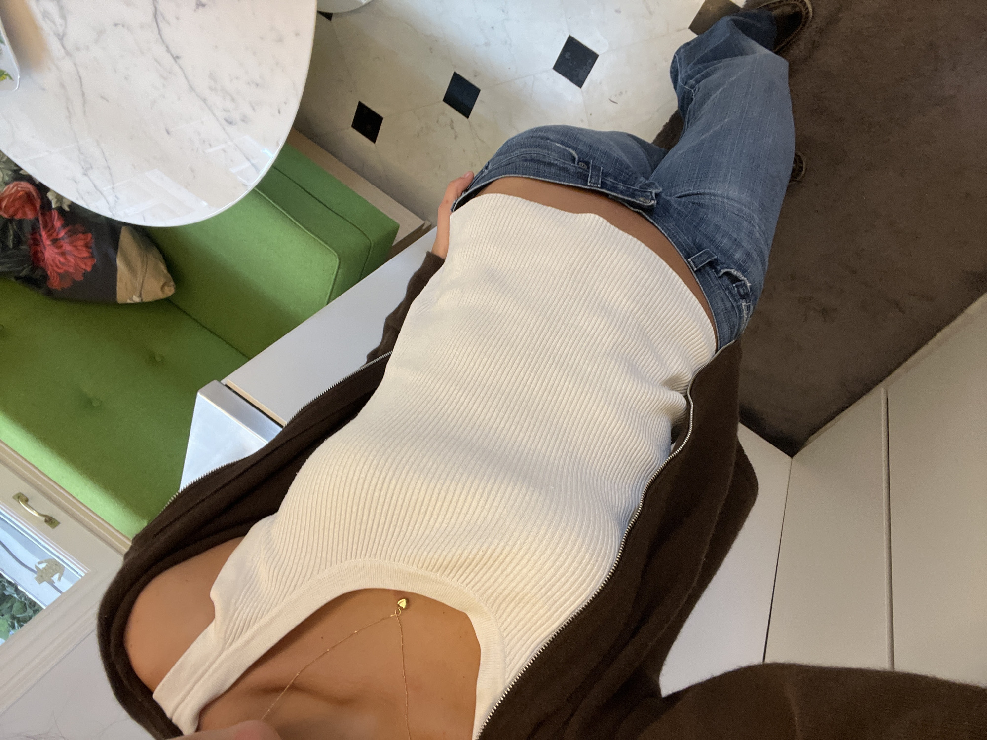 The image provides a high-angle, first-person perspective, looking down at the lower torso and legs of an individual within an indoor setting. The person is wearing a fitted white ribbed tank top, blue denim jeans, and an open dark brown zip-up hoodie. A delicate gold chain necklace with a small heart-shaped pendant hangs around their neck. Dark shoes are visible on the feet.
To the left of the frame, a partial view of a white marble-patterned round table is present, beneath which sits a bright green upholstered bench or sofa. A dark cushion featuring a prominent red floral design rests on the sofa. Further left, a window with white trim and a gold-colored handle shows some greenery outside, indicating natural light.
The floor beneath the person consists of light-colored tiles with distinct black square tiles arranged in a scattered pattern. A dark brown rug or carpet extends under the person's right leg. Along the far right edge of the image, several white vertical panels, possibly cabinet doors, are partially visible.
The overall scene suggests a casual daytime setting, likely a contemporary cafe, kitchen, or dining area, possibly in Paris, France, given the contextual information. The light from the window indicates it is daytime. There is no visible text in the image.