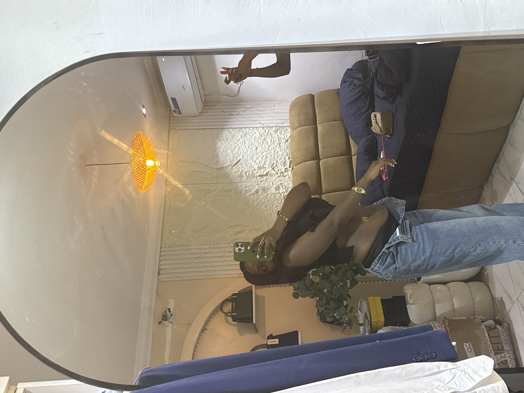 A woman is taking a mirror selfie in what appears to be a bedroom in Abuja, Nigeria. She is wearing a black crop top and light wash jeans. Her hair is dark and long. In the reflection, another person's hands are visible, appearing to hold a phone and possibly take a photo. The room features a large bed with dark blue bedding, a headboard, and a small brown purse on the bed. Other objects in the reflection include clothing hanging on a rack, shelves with handbags, a plant, and a hanging lamp. An air conditioning unit is mounted on the wall.