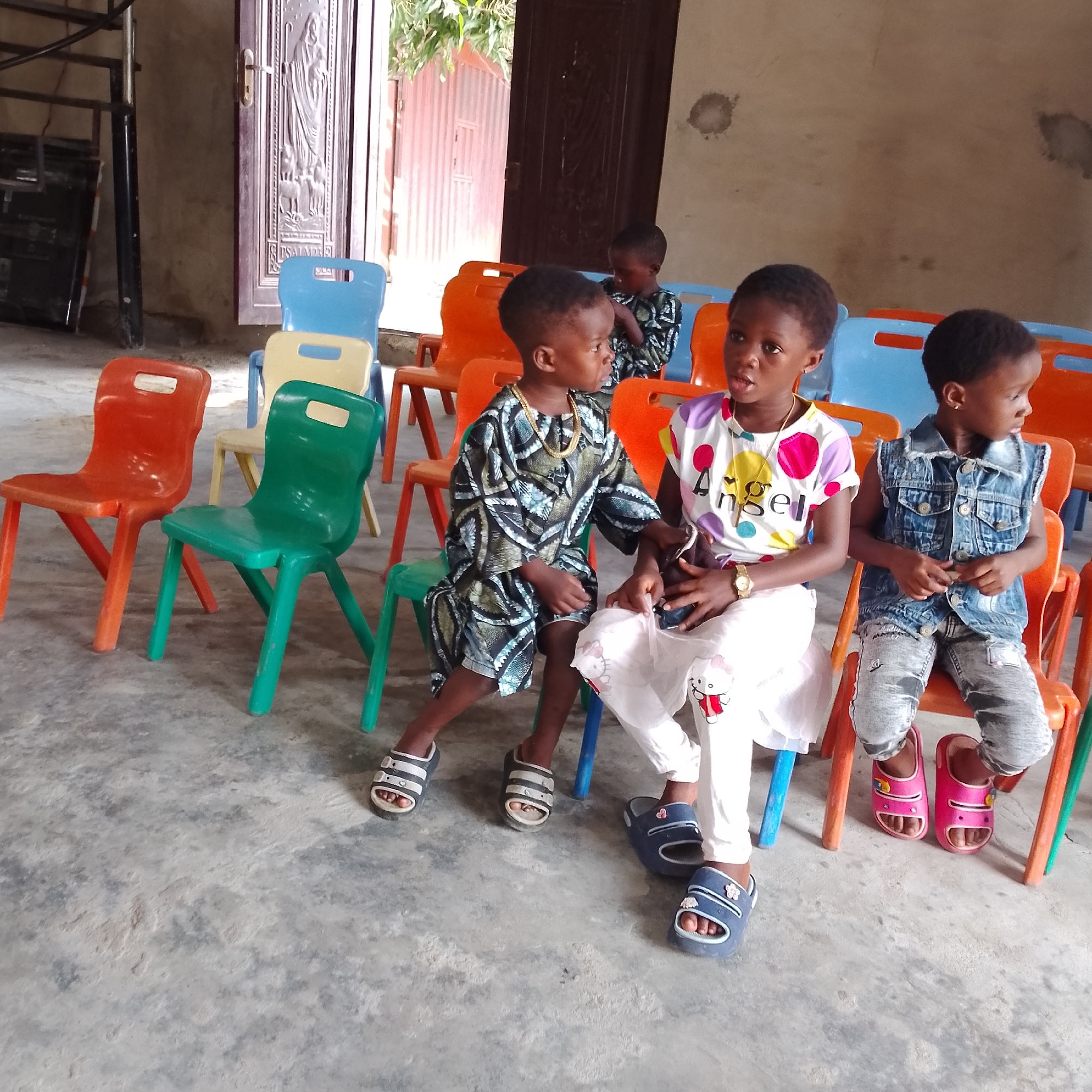 The image depicts four young children seated on colorful plastic chairs arranged in an indoor space, likely a community hall or classroom in Ado, Nigeria. The floor is concrete. The children are positioned towards the right side of the frame, facing away from the viewer, looking to their right.
In the foreground, on the left, a boy is seated on a green chair. He is wearing a traditional dark patterned tunic and shorts, adorned with a prominent gold chain necklace. Next to him, a girl sits on an orange chair. She is wearing a white shirt with colorful polka dots and the word "Angel" printed on it, along with a white skirt. She has a watch on her left wrist. To the right of her, another girl is seated on an orange chair, wearing a denim vest over a white shirt and distressed denim pants. Her feet are in pink slide sandals. The fourth child, partially visible in the background on the far right, is also seated on an orange chair.
The background includes a large metal door with ornate carvings and a handle, likely leading outside. To the left of the door, a metal staircase is visible. In the middle ground, there are several empty plastic chairs of various colors including blue, orange, and yellow. The walls are a neutral, light color, with some circular discolorations. The overall impression is that of a gathering for children, possibly for an event or lesson.