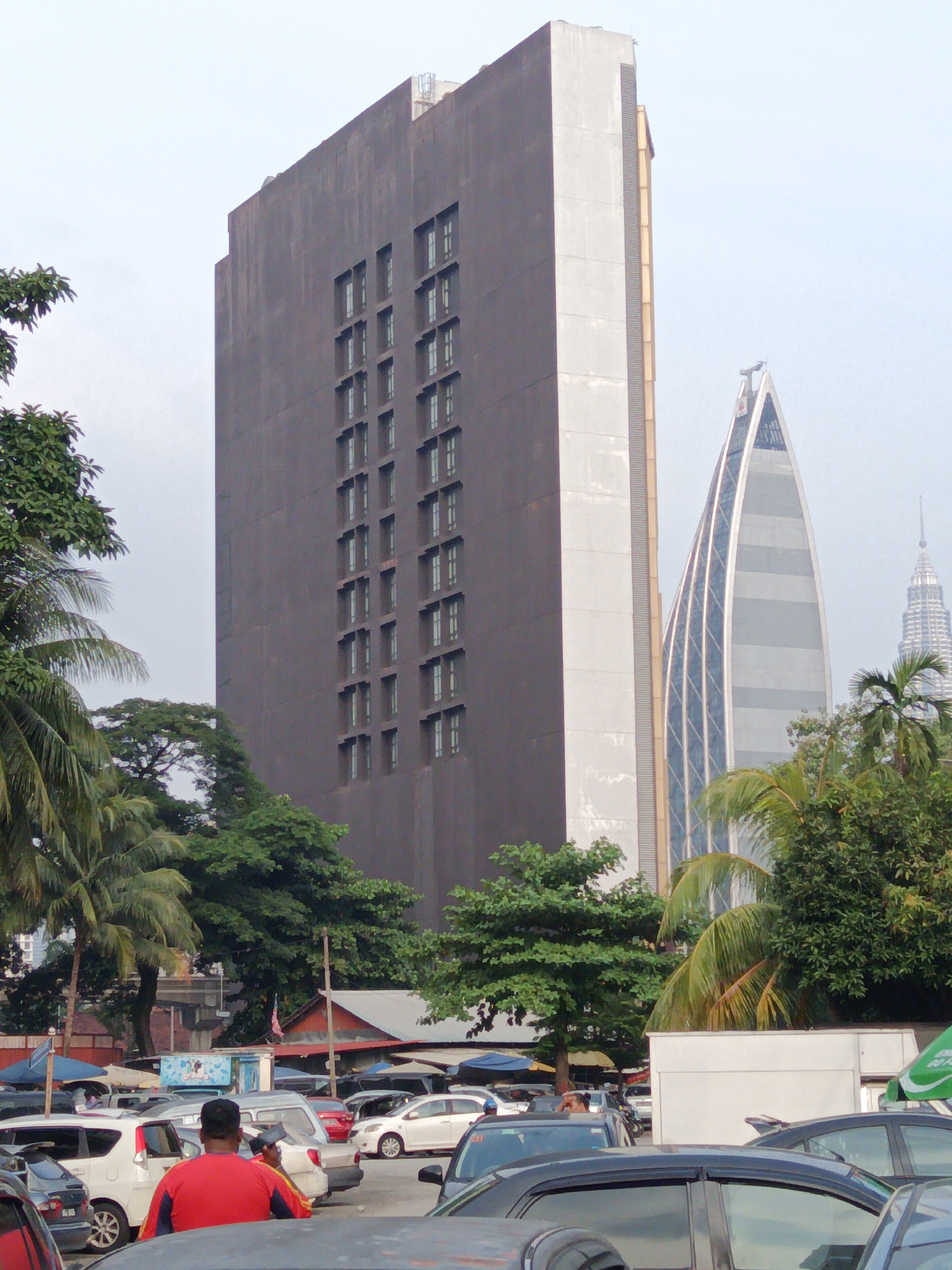 The image shows a street scene in Kuala Lumpur, Malaysia, featuring a distinctive dark rectangular skyscraper dominating the left side of the frame. To the right of this skyscraper, a white, curved, pyramid-shaped building is visible, with the iconic Petronas Twin Towers in the far background. The foreground is filled with parked cars, with a man in a red shirt facing away from the camera, holding a mobile phone. Trees, including palm trees, are scattered throughout the scene, suggesting a tropical urban environment. The sky is overcast.