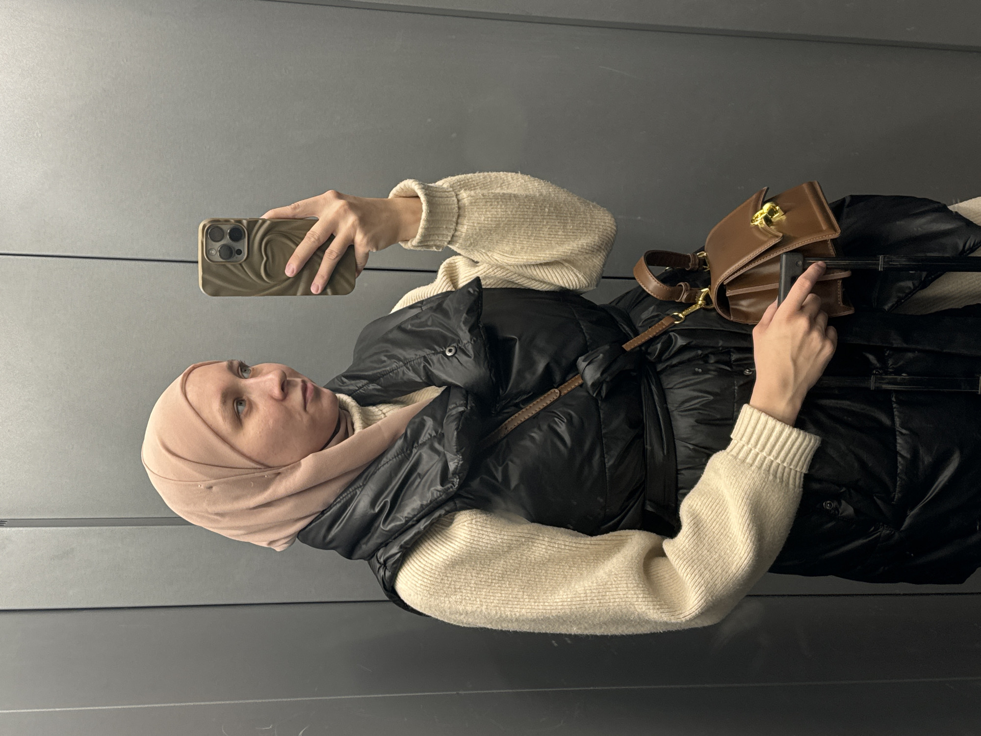 A person wearing a beige hijab, a beige sweater, and a black puffer vest is holding a smartphone in their left hand and a brown leather purse on a suitcase handle with their right hand. The person is standing against a grey wall. The location is not apparent from the image.