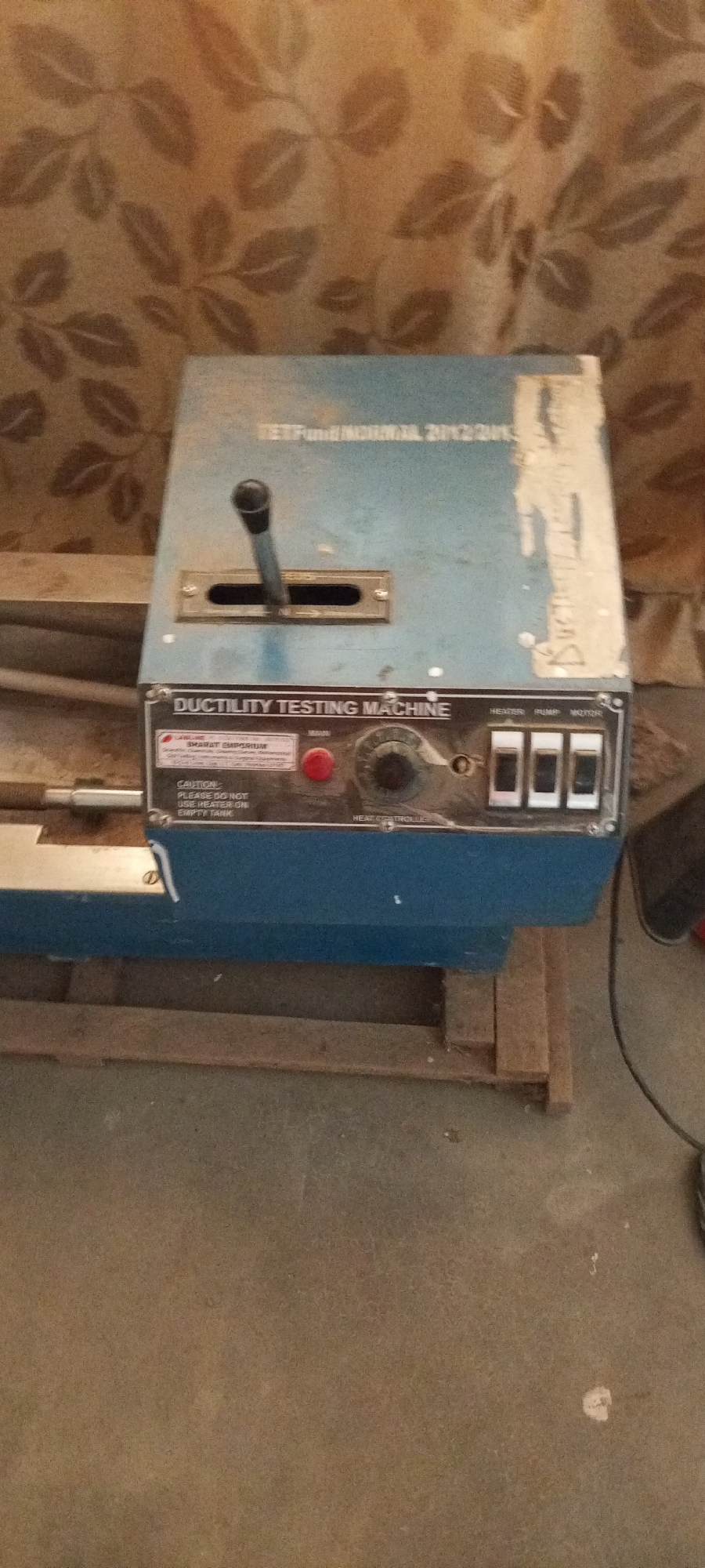 The image shows a blue ductility testing machine with a handle and a dial. The machine is labeled "DUCTILITY TESTING MACHINE" and has several switches on the front. There is also a red button labeled "MAIN". A sticker on the machine reads "BHARAT EMPORIUM" and "CAUTION: PLEASE DO NOT USE HEATER ON EMPTY TANK". The machine appears to be in a workshop or industrial setting, with a brown patterned curtain in the background and a concrete floor. The lighting is dim, and the image is slightly out of focus. There is no indication of the machine's location in Jalingo, Nigeria.