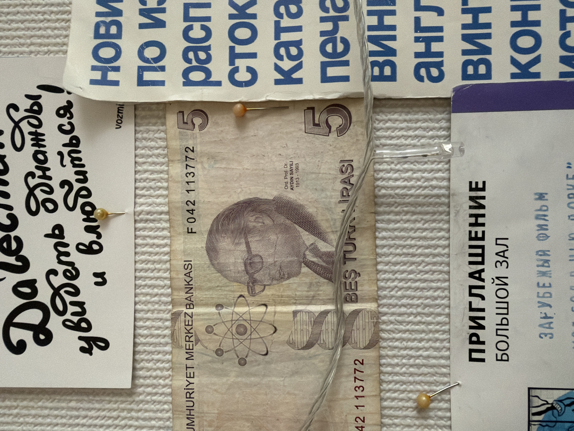 This close-up, flat lay image displays an assortment of personal and informational items pinned onto a white, textured surface, possibly a fabric or corkboard, suggestive of a personal display or mood board.
Central to the composition is a vertically oriented 5 Turkish Lira banknote. The front features the portrait of Ord. Prof. Dr. Aydın Sayılı (1913-1993), a Turkish historian of science. Text visible on the banknote includes "CUMHURİYET MERKEZ BANKASI" (Central Bank of the Republic), "BEŞ TÜRK LİRASI" (Five Turkish Lira), and the serial number "F042 113772". An atom model graphic is also visible next to the portrait.
Several pieces of paper or card with Cyrillic text are pinned around the banknote. To the left, a white card displays black, cursive script, partly visible as "Да... увидеть однажды и влюбиться!" (To... see once and fall in love!). A smaller word "vozmi" (take) is also present.
Above the banknote, multiple strips of paper with blue, block Cyrillic text are arranged, featuring words such as "НОВИ" (New), "ПО ИЗ" (By/From), "РАСП" (likely part of "расписание" - schedule or "распечатать" - print), "СТОК" (Stock), "КАТА" (likely part of "каталог" - catalog), "ПЕЧА" (likely part of "печать" - print/stamp), "ВИН" (Wine/Win), "АНГЛ" (English), "ВИНТ" (Screw/Propeller), and "КОН" (End/Con.).
To the right, a white card with black, block Cyrillic text reads "ПРИГЛАШЕНИЕ" (Invitation), "БОЛЬШОЙ ЗАЛ" (Big Hall), and "ЗАРУБЕЖЫЙ ФИЛЬМ" (Foreign Film), indicating an invitation to a foreign film screening in a large hall.
The items are secured by gold-headed thumbtacks. A thin, clear string or wire crosses over the Turkish banknote, adding another layer to the arrangement. The lighting is even, suggesting an indoor setting without strong directional light or time-of-day cues. The combination of Turkish currency and Cyrillic text from Russia (Zasechnoye) suggests a personal collection or display reflecting diverse interests or travels.