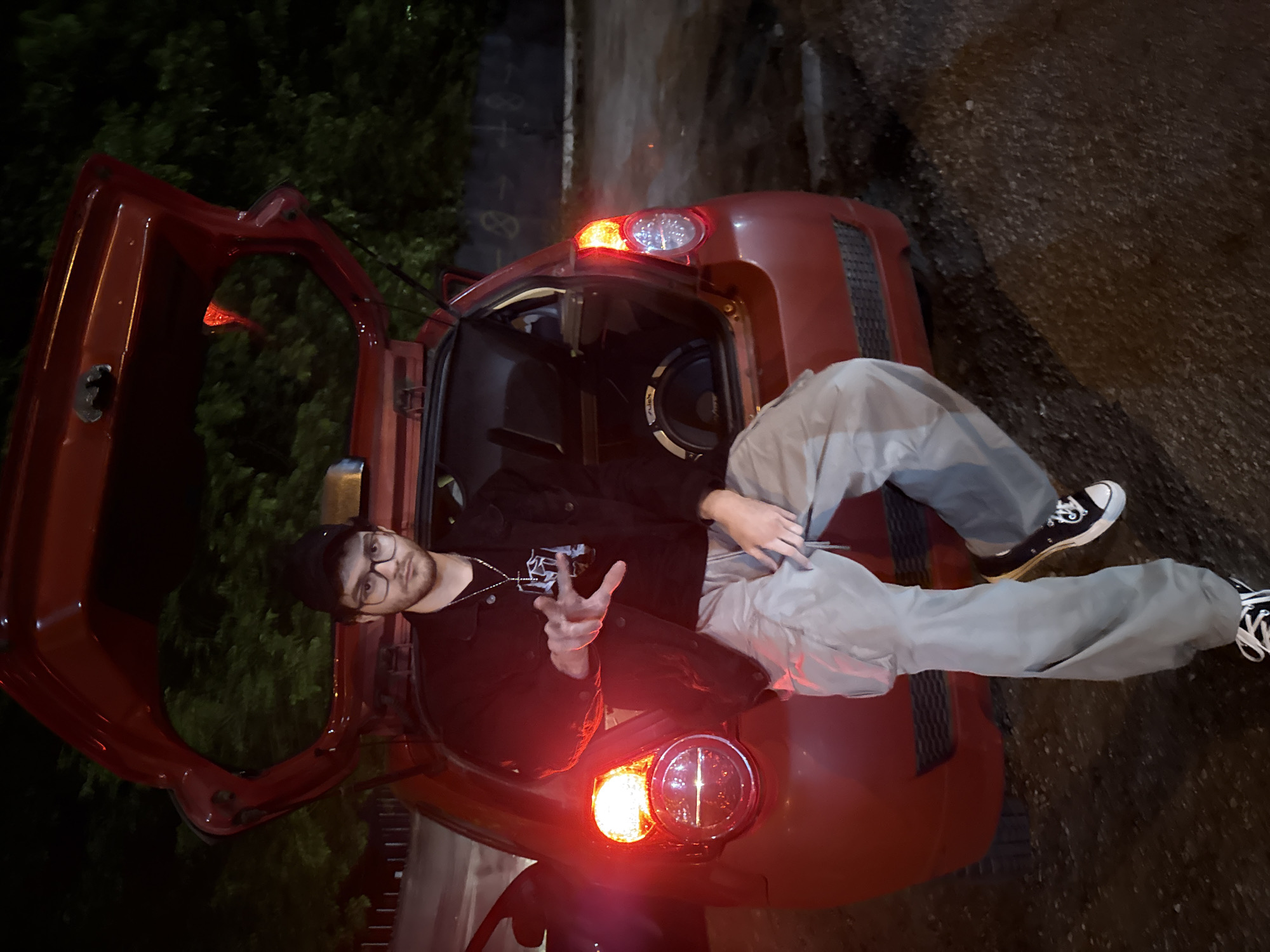 A young man is reclining in the open trunk of a red hatchback car. He wears glasses, a black jacket, a cross necklace, and a dark cap. He also has light gray baggy pants and black and white sneakers. His right hand is raised, making a hand gesture. A large subwoofer is visible inside the trunk. The car's taillights are illuminated. The background suggests an urban environment at night. Although the exact location in Blagoevgrad, Bulgaria, cannot be determined from the image, the setting appears to be a street or parking area, given the presence of the car, streetlight, and vegetation in the background.