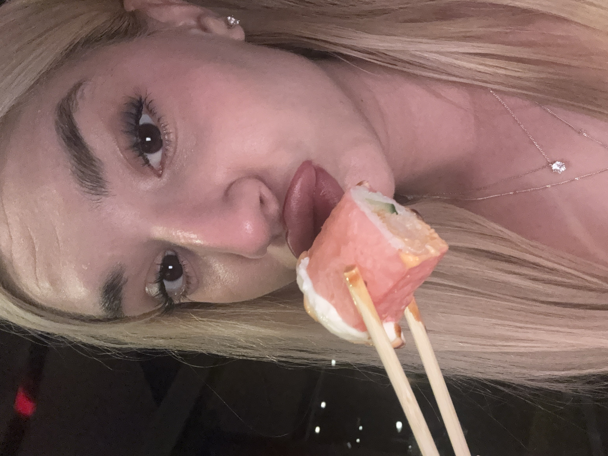 A close-up, rotated portrait shot captures a woman about to eat a piece of sushi. The woman, with fair skin and long, light blonde hair, is positioned on her side, facing the viewer. Her dark brown eyes are open, with visible eyelashes and well-defined eyebrows. Her lips are slightly parted, and her tongue is extended towards the sushi, indicating an imminent bite.
She is holding a vibrant piece of sushi with two light-colored chopsticks. The sushi appears to be wrapped in a thin, reddish-pink fish (likely tuna or salmon), with a creamy white sauce or filling visible on one side and a thin green element, possibly cucumber, peeking out from the top.
The woman wears a delicate, thin silver or white gold necklace with a small, sparkling pendant, likely a diamond, resting on her collarbone. A small diamond stud earring is visible in her upper earlobe. Her hair cascades around her shoulders and back, creating a sense of movement. The background is mostly dark and out of focus, suggesting an indoor setting with low lighting, possibly a restaurant or home environment. The overall mood is playful and focused on the act of eating. No text is visible in the image. The implied location is Sofia, Bulgaria.