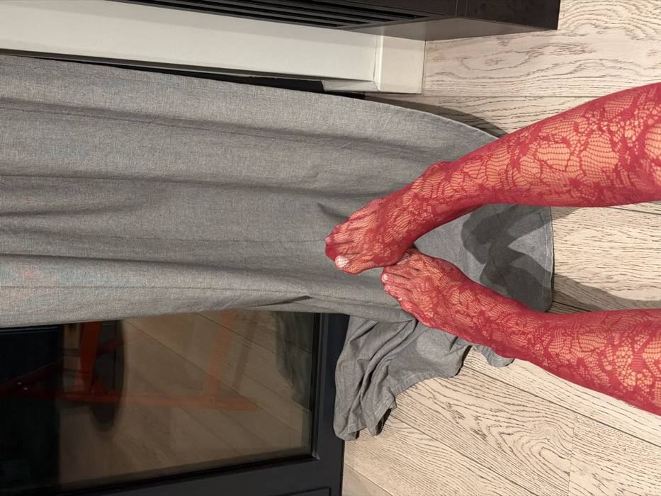 The image captures a close-up, high-angle view of a person's legs and feet adorned in vibrant red, sheer hosiery. The hosiery features an intricate, delicate lace pattern, possibly floral or leaf-like, covering the entire visible length of the legs and feet. The toenails are painted with a clean white polish, providing a subtle contrast to the red.
The legs are positioned with the feet relatively close together, appearing to be resting on a surface. They extend from the bottom right of the frame towards the upper left.
The background elements suggest an indoor setting. To the left of the legs, a substantial piece of light grey fabric, possibly a curtain or drapery, hangs vertically, showing horizontal folds and a slight texture. Below this grey fabric and to the lower left, a dark-framed window or glass door is visible, reflecting indistinct shapes and colors from the interior, including a blurry orange-red object.
Beneath the feet and extending towards the right side of the image is a light-colored wooden floor with a visible grain pattern. At the very top of the frame, a white horizontal surface, likely a window sill or a part of a wall structure, is present, above which a dark, slatted object (possibly a vent or blinds) is situated.
The lighting appears to be ambient indoor light. The photograph seems to be a still shot focused on showcasing the patterned hosiery in a home environment. No specific activity is depicted other than the posing or resting of the legs. No text is visible in the image.