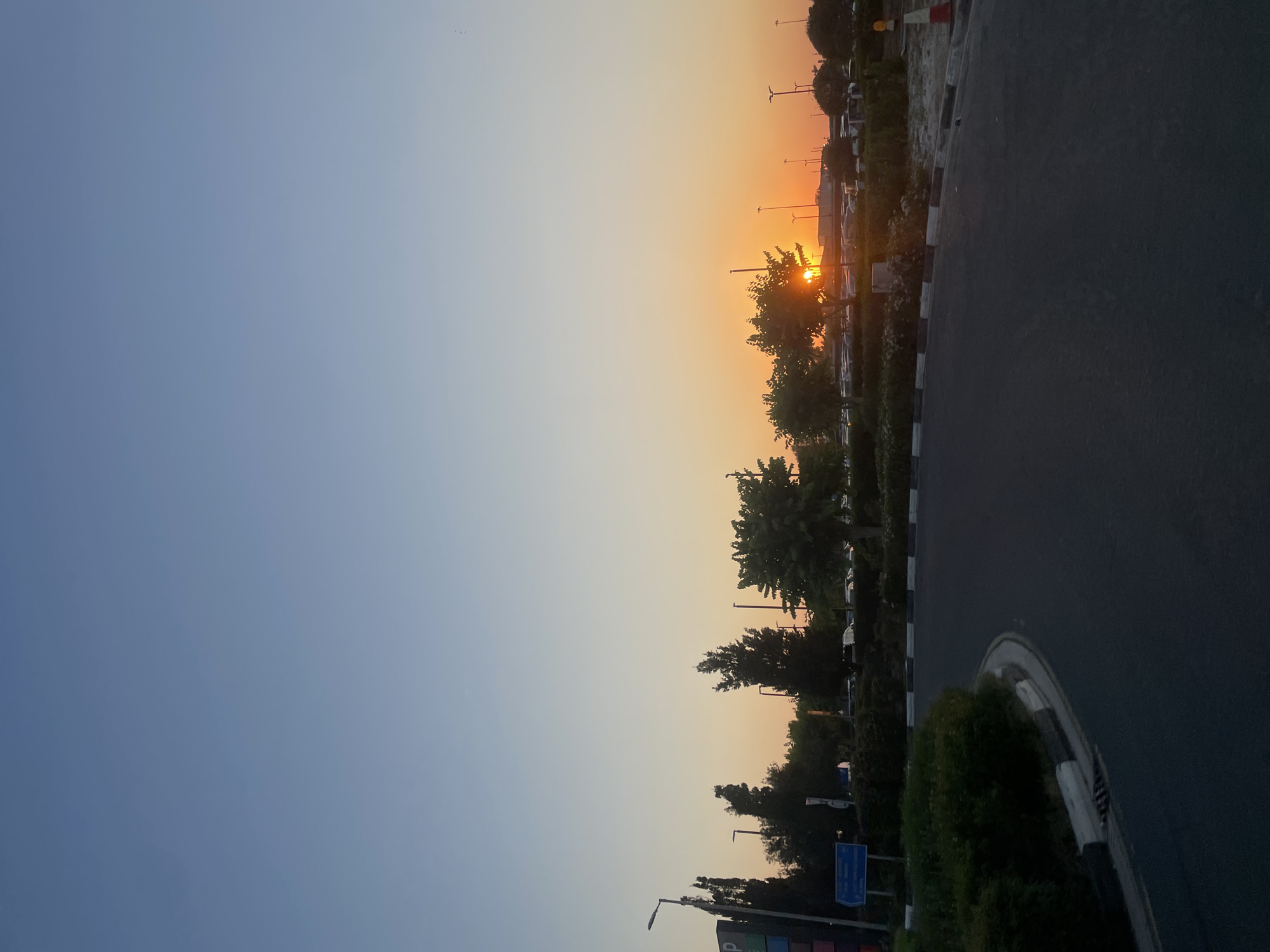 The image shows a clear sky with a gradient of blue to orange, indicating either sunrise or sunset. In the foreground, there is a paved road curving to the left, bordered by a concrete curb with black and white markings. To the left of the road, there is a landscaped area with green bushes and trees. A tall streetlamp stands on the left side of the road, next to a dark signpost with some text that is partially obscured. Further back, beyond the trees and bushes, a parking lot with several cars is visible. The sun is setting or rising behind some trees, casting a warm glow on the sky. In the background, there are more trees, streetlights, and a hint of buildings or structures. The scene appears to be an outdoor public space, possibly near an airport or a commercial area, given the presence of parking and streetlights. The location is identified as Timi Community, Cyprus. There are no people visible in the image.