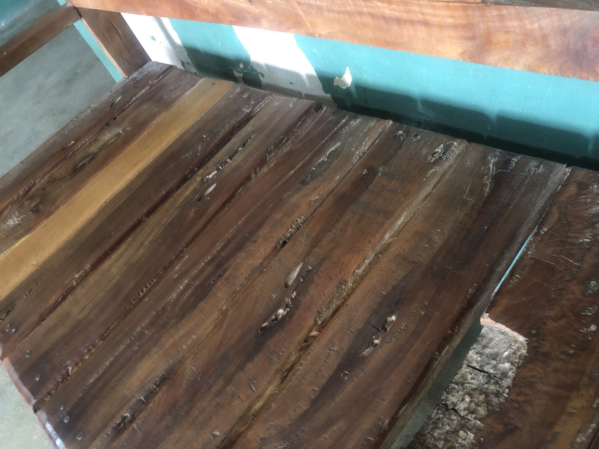 The image shows a close-up view of a weathered wooden surface, likely part of a bench or a table, indoors. The wood is composed of several planks of varying shades of brown and amber, showing signs of age and use with visible grain, cracks, and small indentations. Some nails are present, securing the planks together. To the right of the wooden surface, a vertical turquoise-painted beam is visible, with sections of peeling paint revealing a white underlayer. Beyond this, another piece of wood with a lighter reddish-brown hue is partially visible. The overall impression is of an aged, rustic outdoor or semi-outdoor structure. The specific location in Jalingo, Nigeria, cannot be determined from the visual information provided in the image alone.