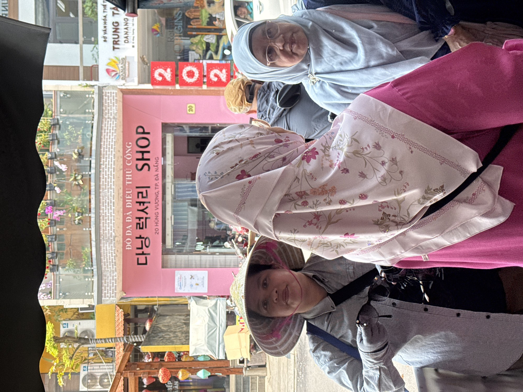 Three women of Asian descent, partially visible, are positioned in the lower right quadrant. One woman wears a traditional conical hat, a light blue shirt with buttons, and holds eyeglasses in a gloved hand. Another woman has a floral-patterned head covering. The third woman wears eyeglasses and a light blue head covering. A pink garment is draped around the women.
Behind the women is a commercial storefront at "20 Hùng Vương, TP. Đà Nẵng". The storefront is painted pink and has text in Vietnamese and Korean ("ĐỒ DA ĐÀ ĐIỂU THỦ CÔNG 다낭 럭셔리 SHOP"). Signage is visible for a cultural center ("SỞ VĂN HÓA, THỂ TH TRUNG TÂ DANANG"). Lanterns hang near the storefront. Air conditioning units are visible on the upper levels of the building. A black object partially obscures the left side of the frame.