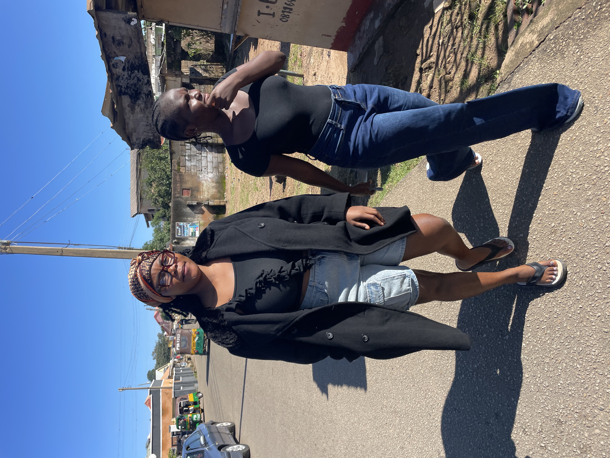Two women are walking down a paved road in Jos, Nigeria, on a sunny day. The woman in the foreground on the left is wearing a black coat, a black ruffled crop top, and light-wash denim shorts with large front pockets. She has on glasses and a patterned headscarf. She is wearing flip-flops. The woman on the right is wearing a black scoop-neck t-shirt and dark wash flared jeans. She has her hair in braids and is wearing sneakers. The road is lined with buildings and trees. In the background, there are several vehicles, including a car, and at least two motorized tricycles, known locally as "keke napeps." The sky is clear and blue. The overall scene is consistent with a street in an urban area of Nigeria.