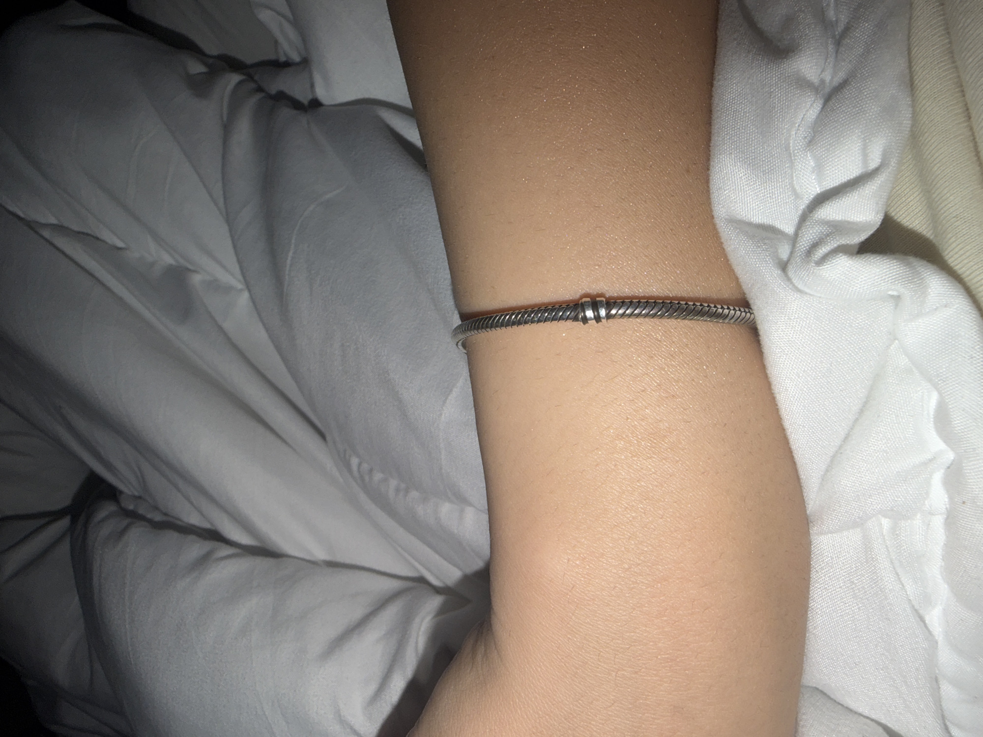 A close-up shot focuses on a person's light-skinned arm, adorned with a silver-colored bracelet, resting amidst white fabric. The arm, positioned centrally in the frame, shows subtle skin texture and fine hair. The bracelet has a snake chain design with two small, plain silver spacer beads near the center.
The background consists of soft, wrinkled white fabric, likely bedding such as sheets or a duvet cover, which appears to envelop or surround the arm. The lighting is dim, suggesting it might be nighttime or a poorly lit room, casting soft shadows and giving the skin a slightly luminous appearance. There is no visible activity other than the arm resting. No text is visible in the image. The overall impression is one of a quiet, intimate moment, possibly in bed.
