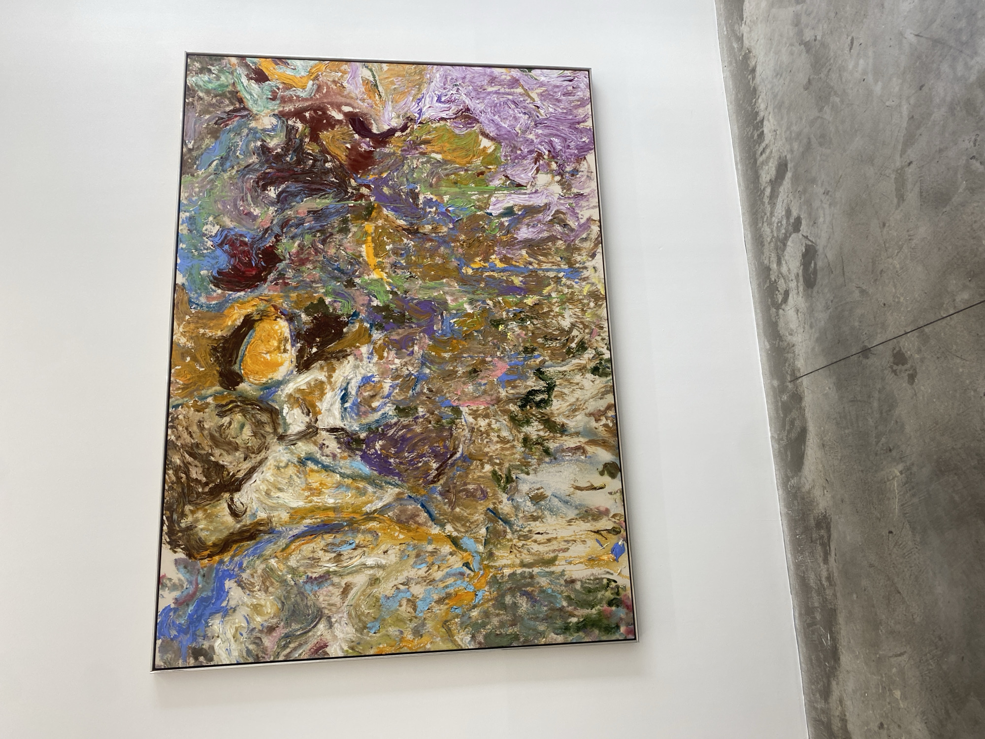A framed abstract painting hangs on a white wall. The painting contains a variety of colors including purple, brown, blue, yellow, and green. The painting is hung near a gray wall. The location is unspecified, but is presumed to be in Paris, France.