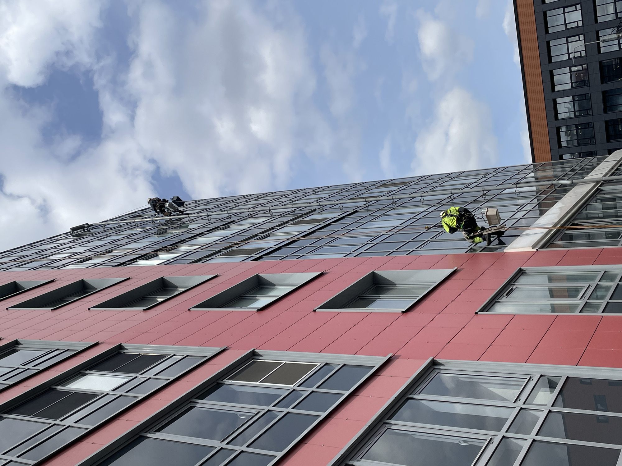 A low-angle shot depicts the facade of a modern building with a complex design, potentially located in Saint Petersburg, Russia. The building has two distinct sections: a lower part with a red, tile-like exterior and an upper part consisting of many mirrored windows. Two window cleaners are visible on the mirrored portion of the building, suspended by ropes. One is wearing a bright green safety vest and appears to be actively working. The sky is visible above the building, filled with puffy white clouds. Another building with dark windows is visible on the right side of the image, adjacent to the main building.