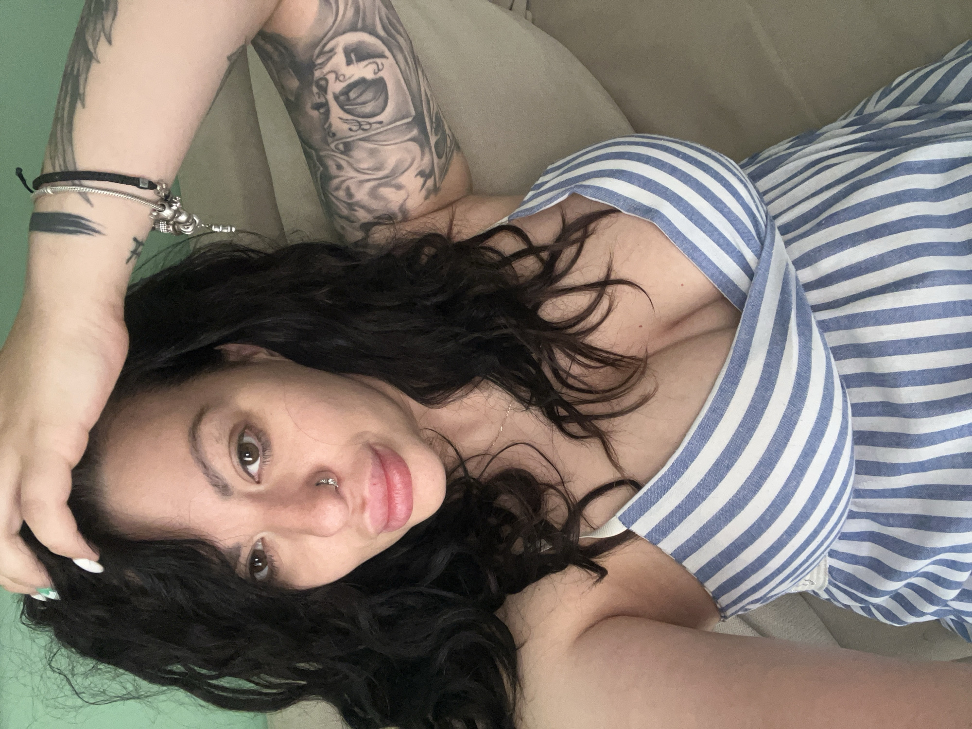 A woman with dark, curly hair and light skin is lying down, possibly on a couch or bed. She is wearing a blue and white striped top with a low neckline. Her left arm is raised above her head, revealing a bracelet and tattoos on her forearm and upper arm. She has a nose ring. The location in Plovdiv, Bulgaria is unknown.