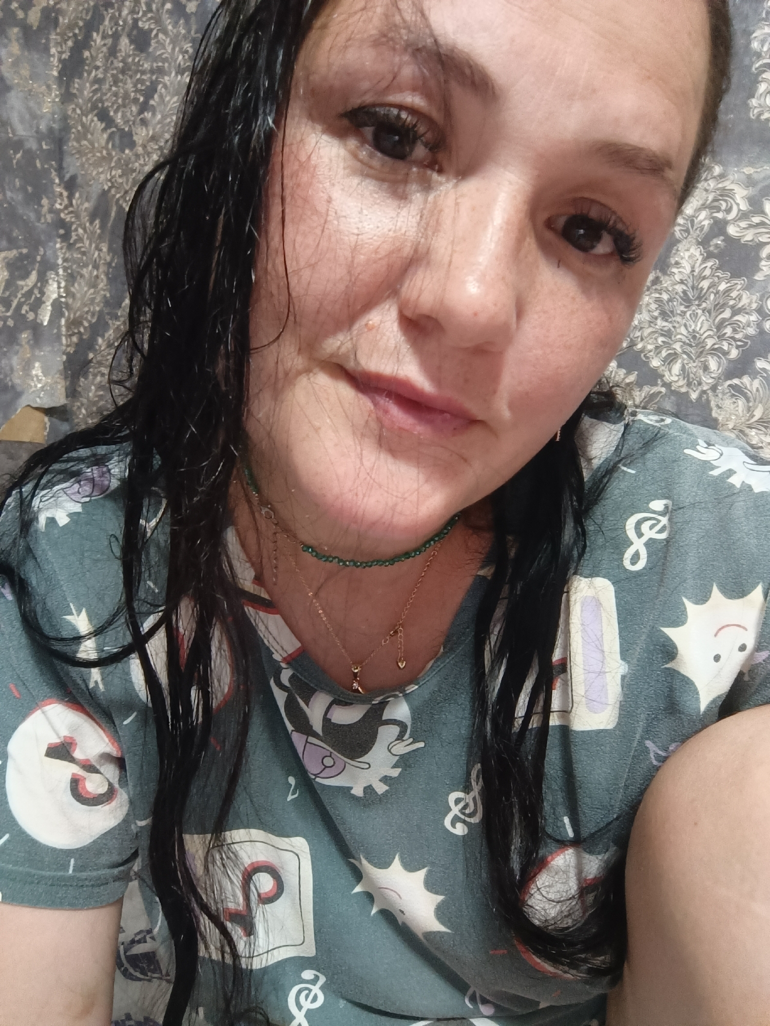 A woman with dark hair and light skin is shown from the chest up, looking at the camera. She is wearing a green shirt with a white pattern of cartoonish figures. A green beaded necklace and a gold chain with a heart pendant are visible. Her hair is dark, wet, and falls around her face and shoulders. She has visible freckles and appears to be in a domestic setting, with a patterned wallpaper in the background. The image was taken indoors.