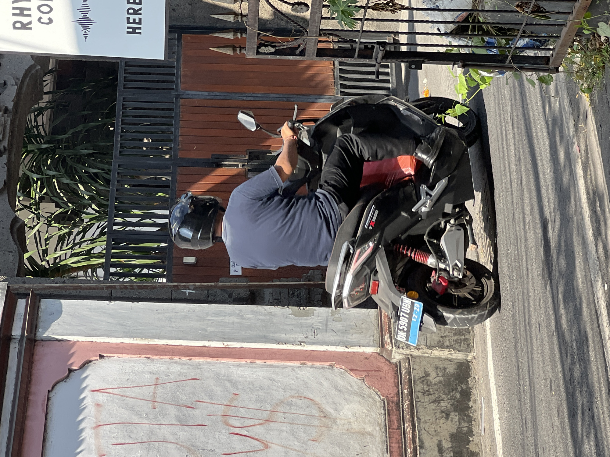 In Kuta, Indonesia, a person is riding a black scooter on an asphalt road, casting a shadow. The scooter has a red rear shock absorber and a blue license plate that reads "DK 5907 UBN 12-29". The rider is wearing a black helmet, a dark blue t-shirt, and black pants. They are holding onto the handlebars with their right hand. To the left of the road, a pink and white wall with red graffiti is visible. To the right, a black metal fence with decorative elements stands in front of a wooden gate and lush green plants. Behind the fence, a building with a sign that says "RHYTHM CO" and "HERE" is partially visible.