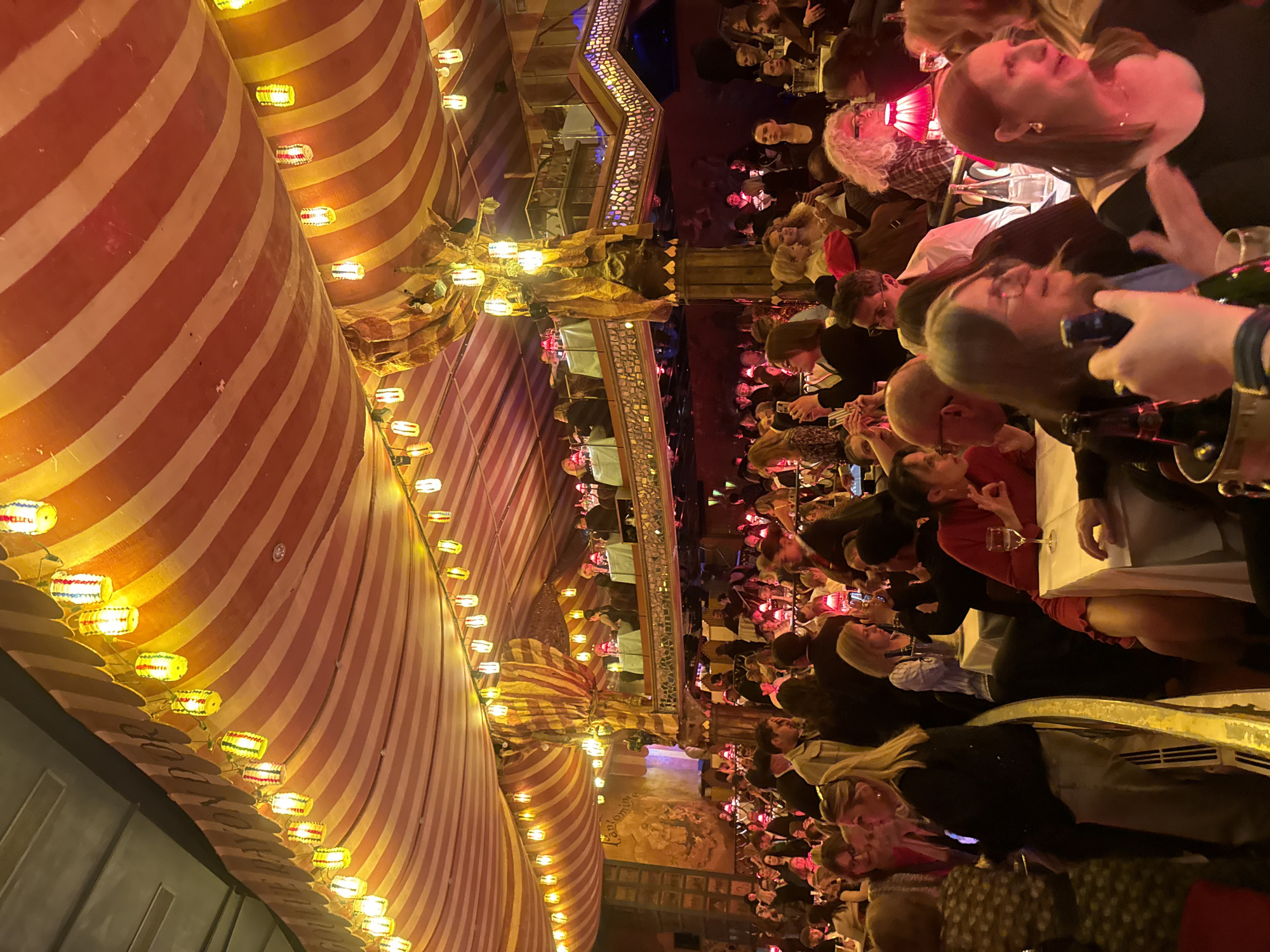 The image depicts a crowded indoor scene, possibly a restaurant or cabaret, likely located in Paris, France, as suggested by the architectural style. Numerous people are seated at tables, appearing to be dining or watching a performance. The atmosphere seems lively, with individuals interacting and engaged with their surroundings.
The interior features a striped ceiling adorned with strings of lights, creating a festive ambiance. There is a balcony or upper level visible, also occupied by patrons. The lighting is warm, contributing to the overall vibrant setting.
Details observed among the crowd include various hairstyles, attire, and visible interactions. Some individuals are using mobile phones, possibly capturing the event or scene. Tables are set with white tablecloths and glasses, indicating dining arrangements.
