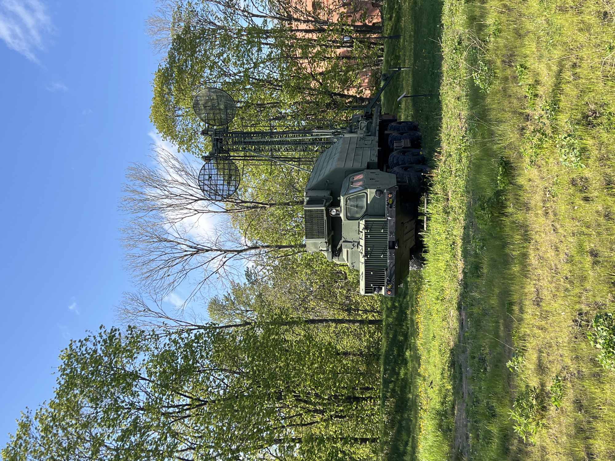 The image depicts a P-19 "Danube" radar system mounted on a truck chassis. The radar system is painted in a camouflage green color. It is positioned on a grassy field, with trees in the background. The sky is partially visible with some clouds. The image was taken in Saint Petersburg, Russia.
