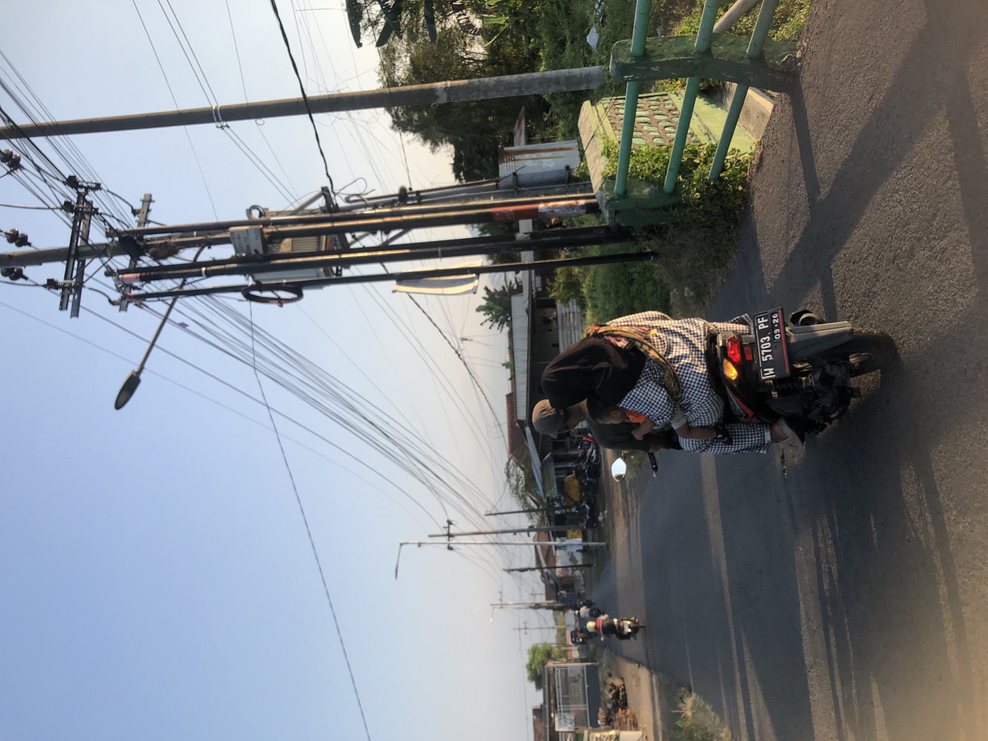 In the city of Gempolsari, Indonesia, this image captures a street scene with a clear blue sky and power lines crisscrossing overhead. In the foreground, a couple and a child ride on a motorcycle. The person in the back, wearing a hijab, holds the child. The motorcycle has a license plate that reads "W 5703 PF" and "09-26". The road is paved, and on the right side of the image, there is a green metal fence overgrown with plants, and a concrete structure. On the left side of the road, there are houses and other buildings. In the distance, other motorcycles are visible on the road. The scene appears to be during sunset, as indicated by the long shadows cast on the road.