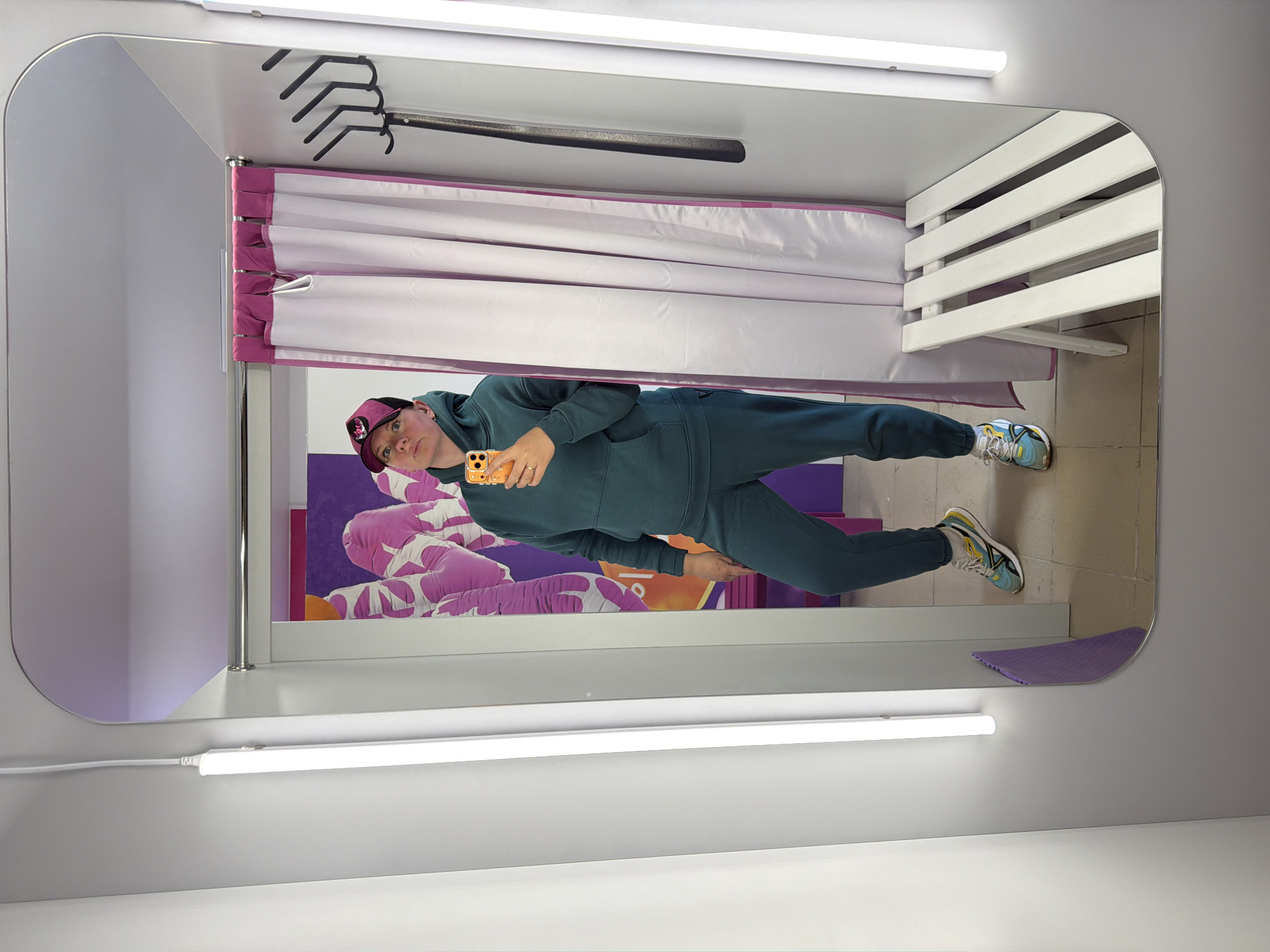 The image is a selfie of a person in a dressing room, likely in Chaikovsky, Russia. The person is wearing a dark green tracksuit, a cap with a logo, and sneakers. They are holding an orange cell phone, presumably taking the picture. The dressing room has a white bench with horizontal slats, a clothes rack with black hangers, and a purple and white curtain. There is a purple backdrop or artwork on the wall behind the person. The room is lit by fluorescent tube lights. A purple cleaning cloth is visible on the floor. The image appears to be taken in a mirror, hence the reversed perspective.