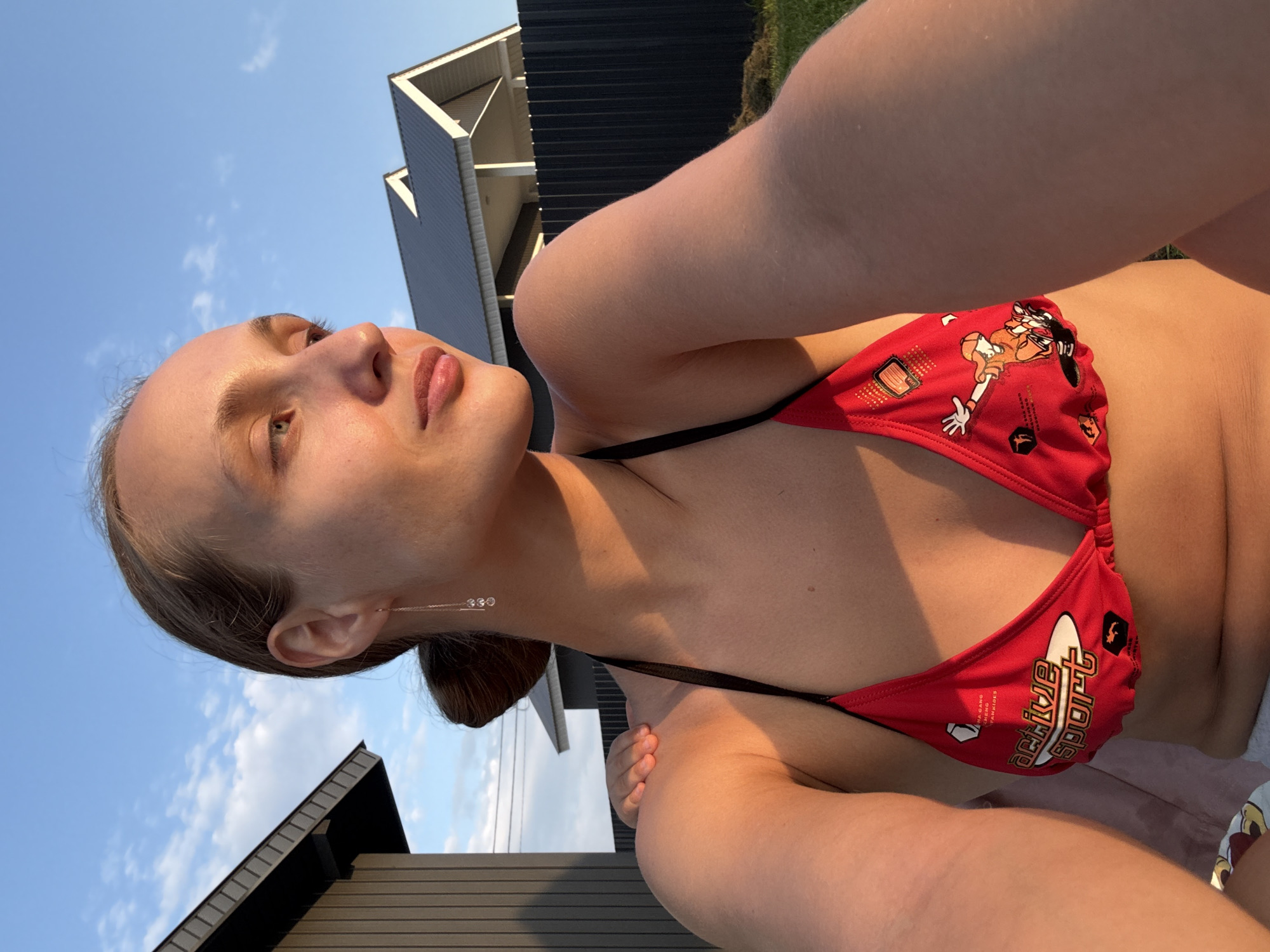 The image depicts a young woman in a red bikini outdoors, with a house and fence in the background. The sky is visible with some clouds. The woman's face is in profile, looking to the right, and her arm is resting on her shoulder. Sunlight is casting a warm glow on her skin. The scene appears to be set in a residential area.