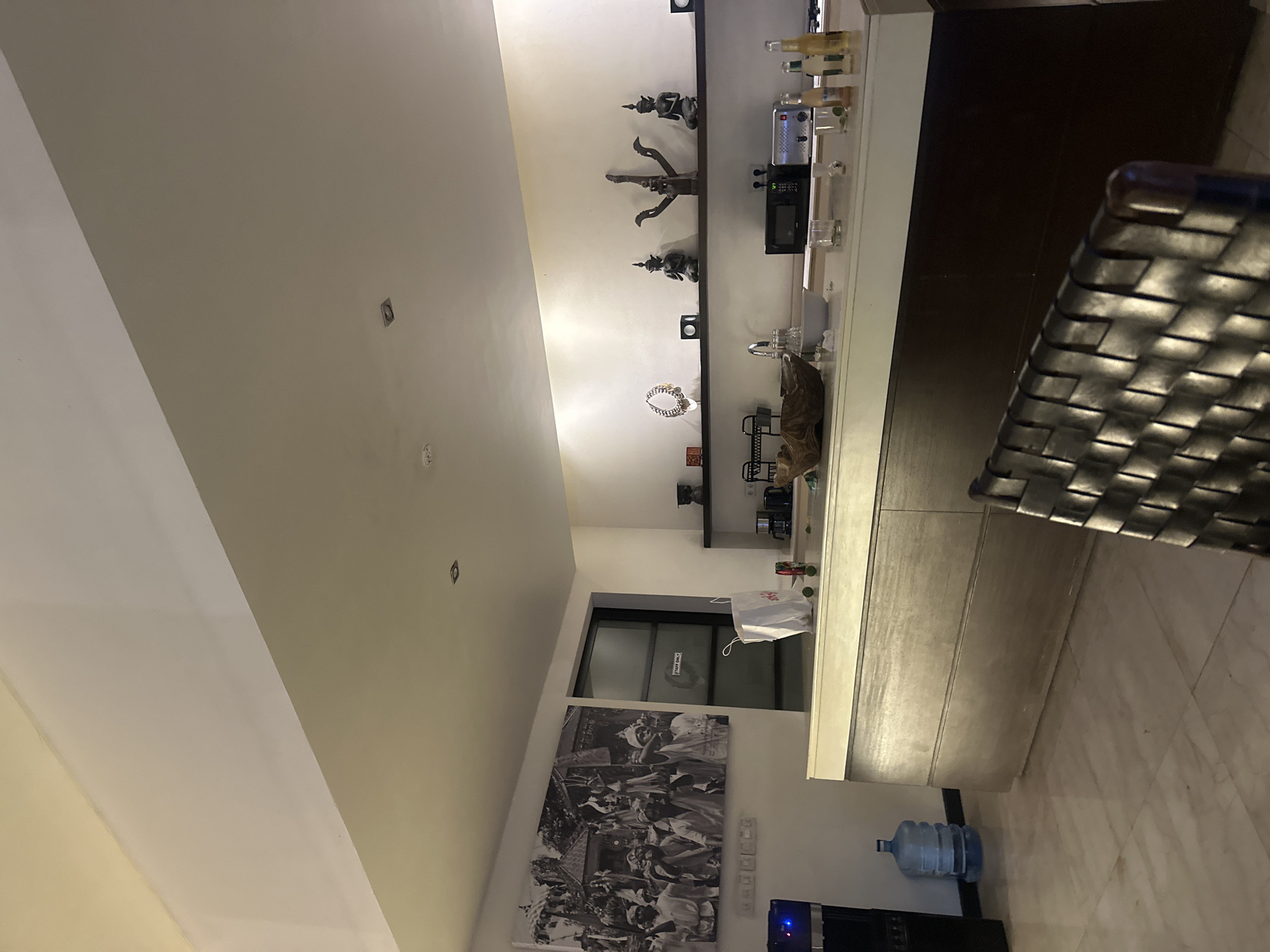 The image shows a kitchen area, likely in North Kuta, Indonesia. The space features a long countertop with a mix of decorative items and appliances. Wall decorations include statues, a photograph, and a black and white print. A water cooler is placed below the counter. A chair is partially visible in the foreground. The lighting is bright, illuminating the white walls and countertop.