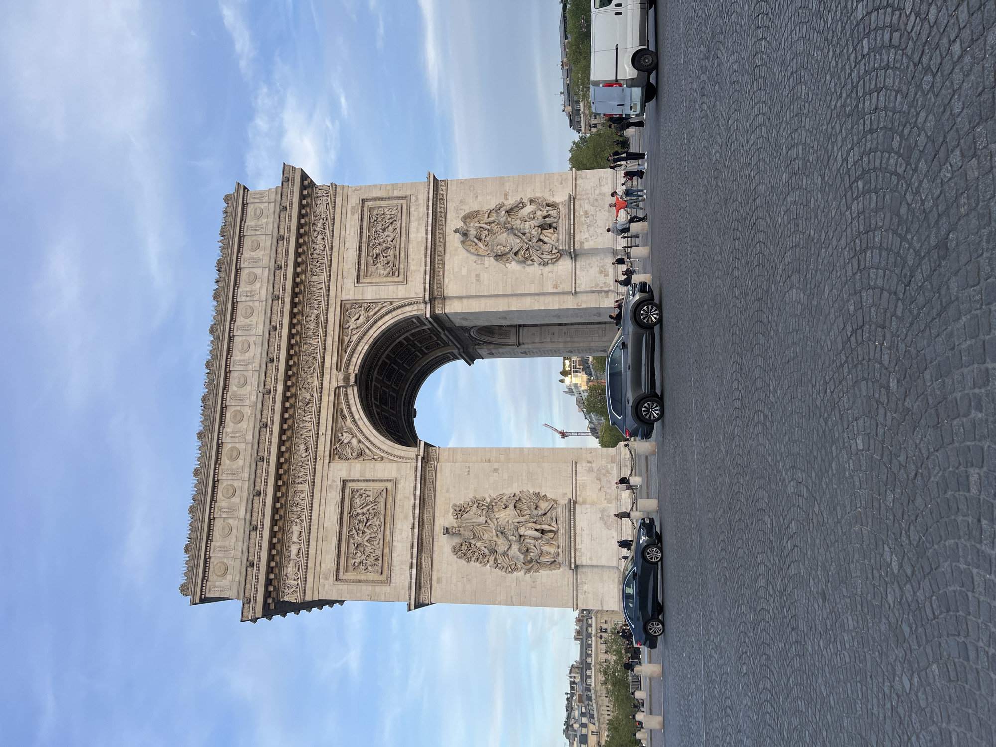 This image, correctly oriented, displays the Arc de Triomphe in Paris, France, against a clear blue sky with delicate, wispy clouds. The grand, light-colored stone monument occupies the left side of the frame, showcasing its elaborate neoclassical architecture. Detailed bas-relief sculptures and ornamental carvings adorn its surfaces; notably, the famous "Departure of the Volunteers of 1792" (La Marseillaise) sculptural group is prominent on one of its pillars.
To the right of the monument, a wide, textured cobblestone road, typical of Paris, stretches into the distance. Several vehicles are visible on or alongside this road: a dark grey sedan is positioned closer to the foreground, another dark car is further back, and a white van is parked towards the upper right.
Numerous people are seen around the base of the Arc de Triomphe and on the sidewalk. They appear to be visitors or tourists, observing the monument and moving about. The scene suggests a busy daytime atmosphere. In the background, behind the monument and road, characteristic Parisian buildings and scattered trees are discernible under the bright, clear sky, indicating pleasant weather. No discernible text is visible on the monument or in the scene.