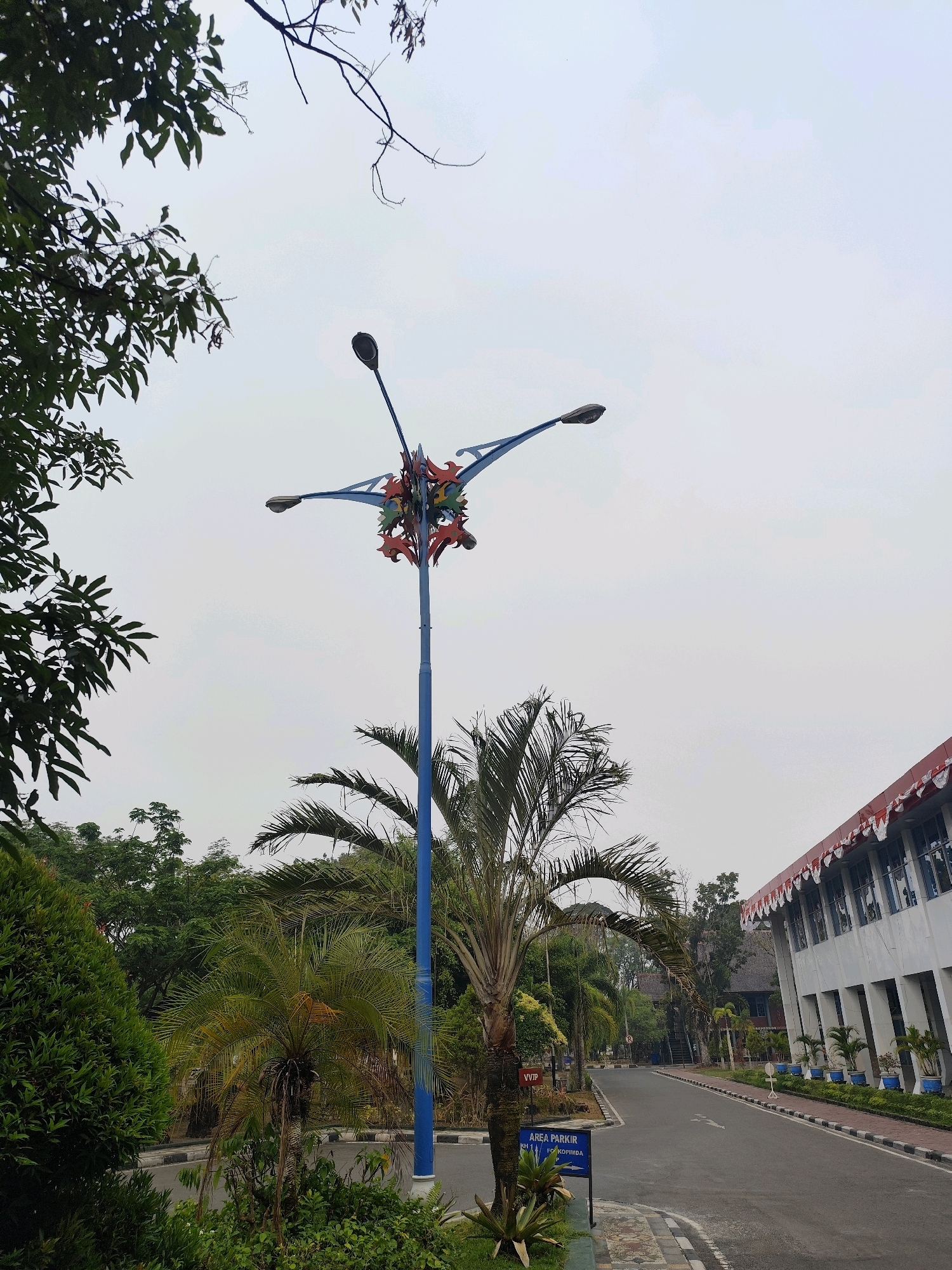 The image depicts an outdoor scene with a street leading to a modern building. A tall, blue lamppost with decorative elements at the top stands prominently in the foreground, partially obscured by trees and foliage. The lamppost has multiple arms, each supporting a light fixture. To the right of the lamppost, a paved road curves away, flanked by a sidewalk and a white building with a red and white decorative awning. The building has numerous windows and is lined with potted plants. Various types of trees, including palm trees, are visible throughout the scene, suggesting a tropical or subtropical environment. A sign indicating "AREA PARKIR" (parking area) is present near the base of the lamppost. The sky is overcast, casting a diffused light on the scene. The overall impression is one of an institutional or public space within a well-maintained green area. The context provided indicates the location is Palangka Raya, Indonesia.