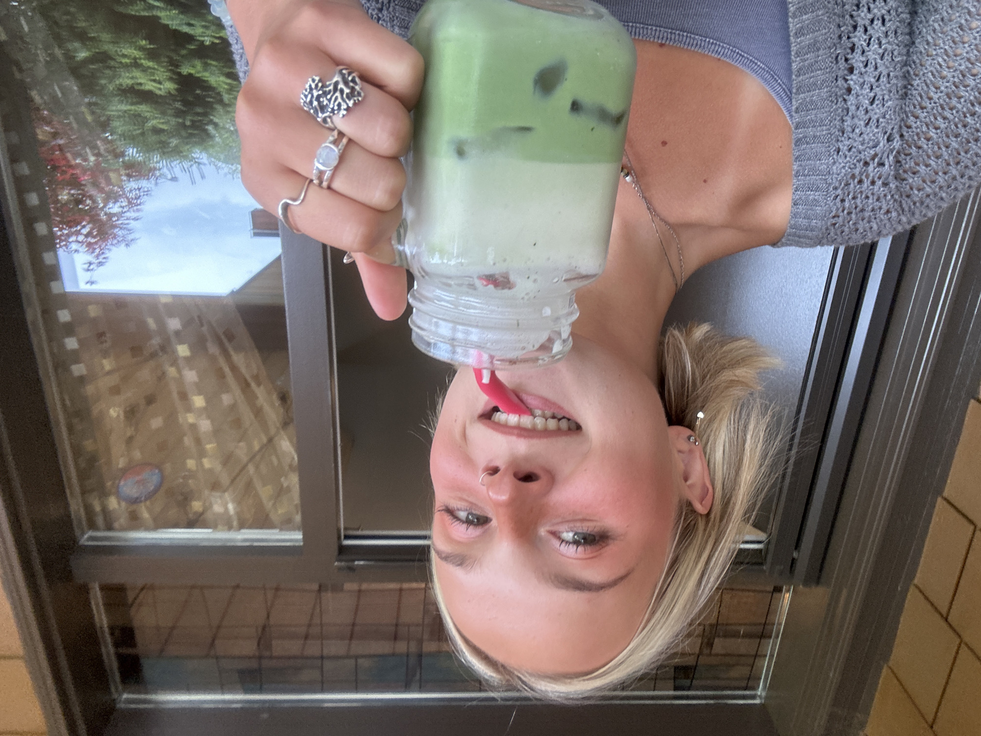 In Whistler, Canada, a young woman with blonde hair and fair skin takes a selfie while holding a glass mason jar filled with a layered green beverage and ice. She is wearing a grey crocheted cardigan over a grey tank top and multiple silver rings on her fingers, one featuring a moonstone and another with a tree-like design. She has a nose ring and her ears are adorned with small earrings. The background suggests an outdoor setting, possibly a patio or porch, with a glass-paneled door and a glimpse of trees and sky. She appears to be drinking from the jar through a pink straw.