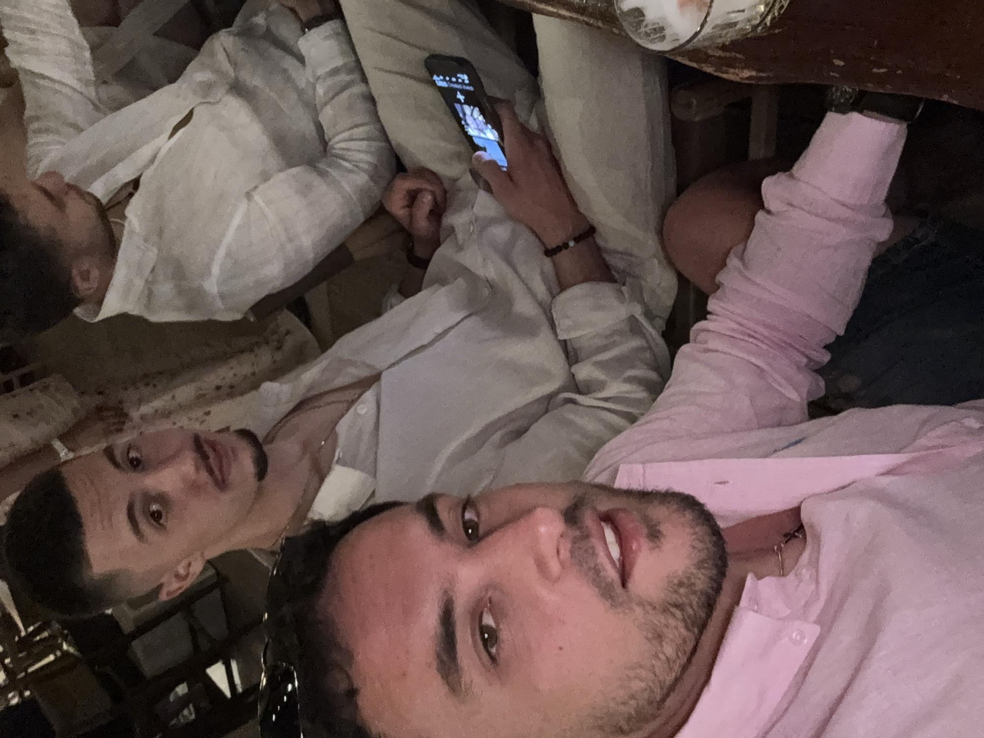 Three men are seated at a table, taking a selfie. One man wears a pink shirt and is positioned closest to the camera. Another man in a white shirt looks at the camera. A third man in a white shirt looks at a cell phone he is holding. The location appears to be indoors, based on the lighting. The location within the city of Pallini Municipal Unit, Greece, is not discernible from the image.