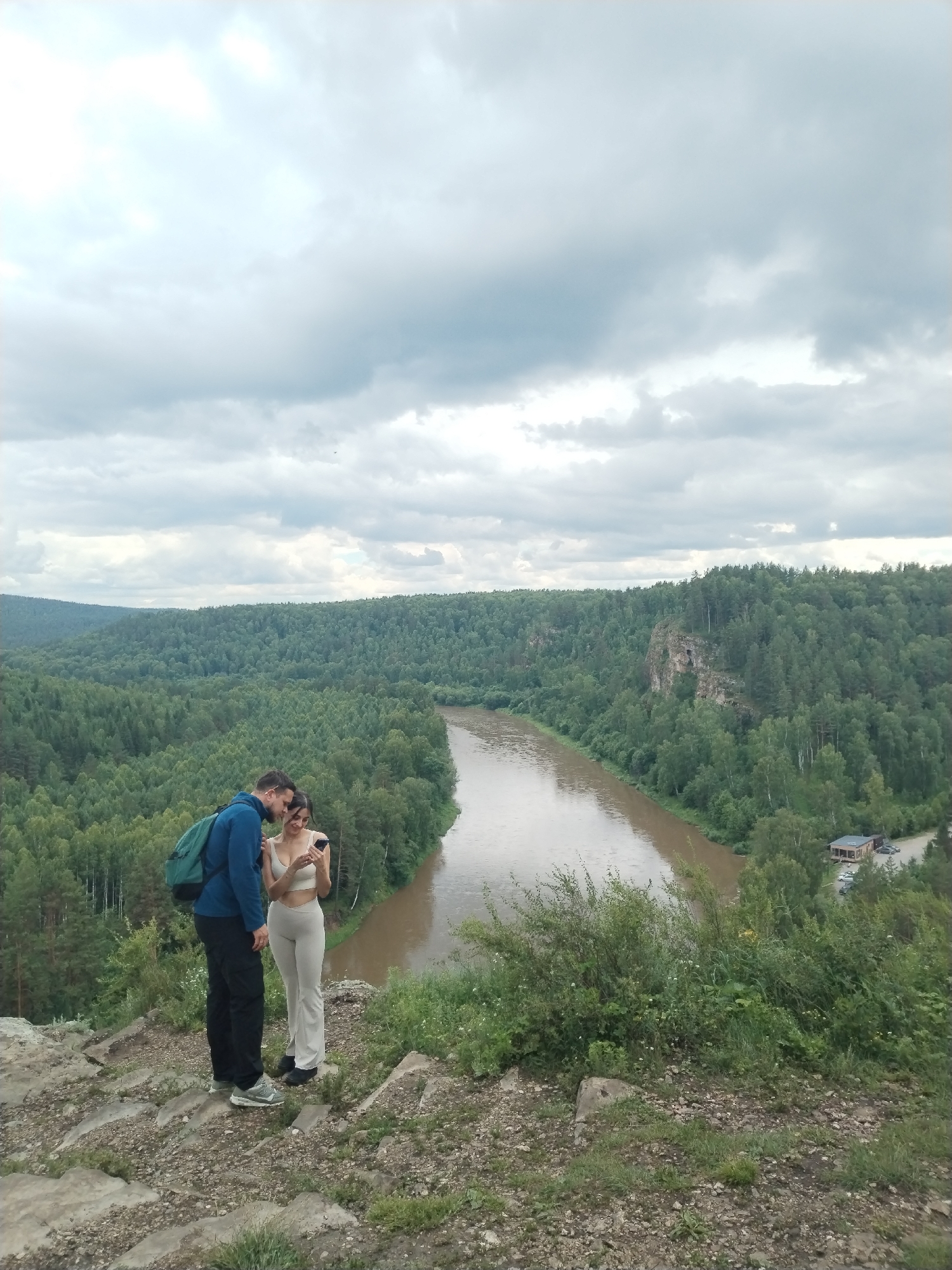 A man and a woman are standing on a rocky incline overlooking a wide, muddy river that curves through a densely forested valley. The man, wearing a blue jacket and black pants, has a green backpack slung over one shoulder. The woman, in a light-colored top and matching pants, is looking at a phone. They appear to be engaged in conversation. The sky is overcast with dramatic clouds. In the distance, a few buildings are visible near the riverbank, and the surrounding hills are covered in a thick carpet of green trees. The scene is likely set in the natural landscape near Idrisovo, Russia, known for its picturesque river valleys and forests.