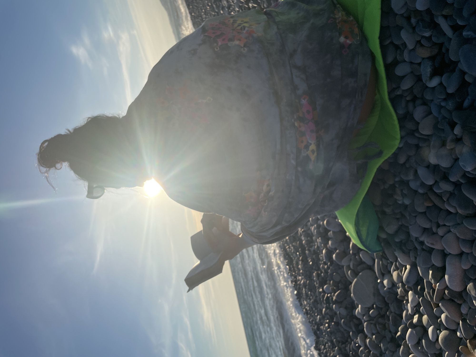 The image shows a person sitting on a pebble beach, facing the ocean. The sun is setting, casting a warm glow on the scene and creating a lens flare effect. The person is wearing a floral-patterned dress and has their hair tied up. They are holding a book and appear to be reading or looking at it. The ocean waves are gently lapping at the shore. The overall mood is peaceful and serene. The time of day is late afternoon or early evening. The weather appears to be clear and sunny. The location is a beach with a pebble shore. There is no visible text in the image.