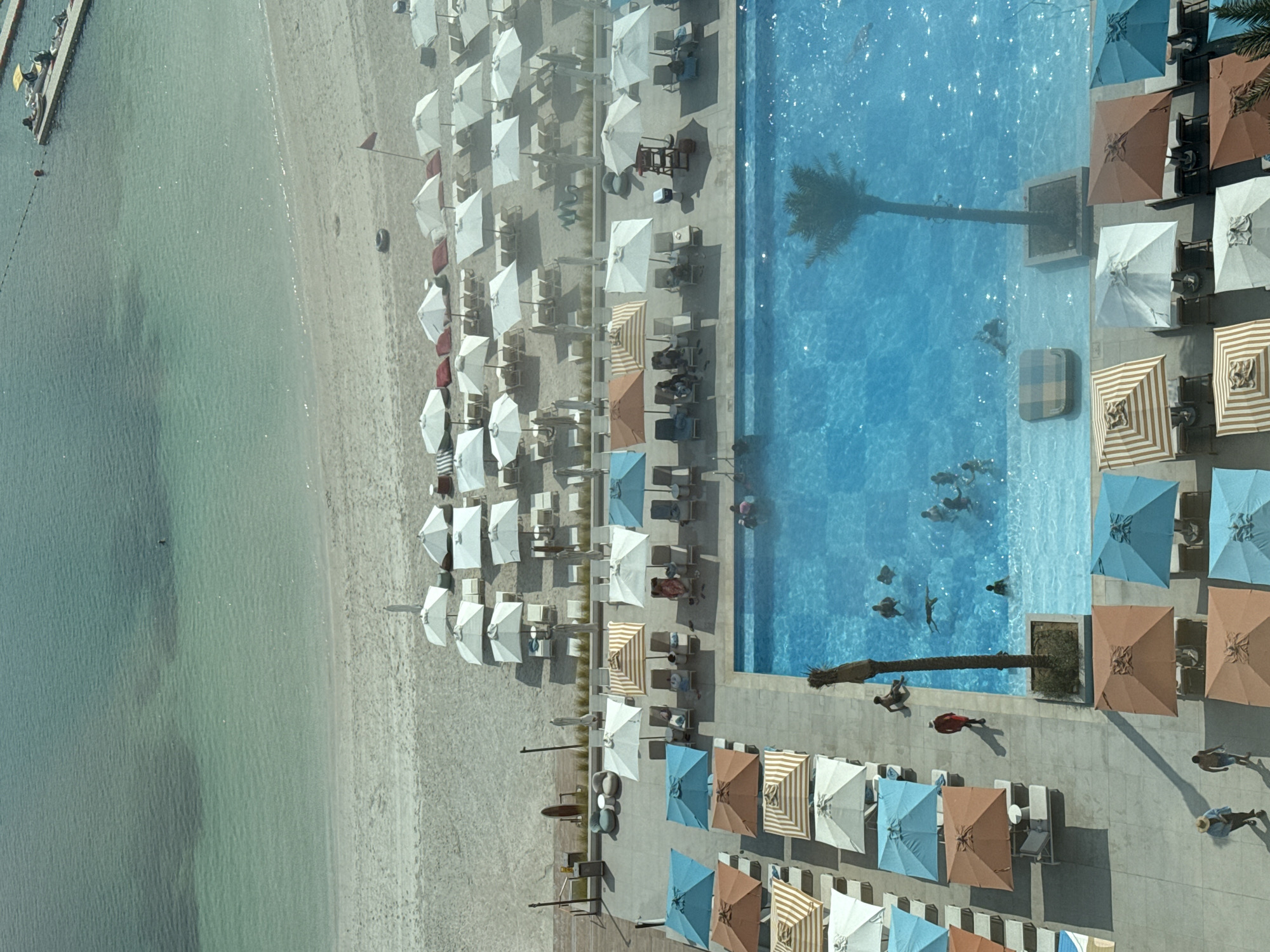 An aerial view shows a resort scene in Ras Al-Khaimah, United Arab Emirates, featuring a sandy beach, a swimming pool, and numerous umbrellas and lounge chairs. The turquoise water of the sea meets the light-colored sand of the beach. Beyond the beach, rows of white umbrellas and lounge chairs are set up. Closer to the foreground, a large, bright blue swimming pool is visible, with several people swimming and relaxing in and around it. Palm trees are also present within the resort area. The scene is bathed in bright daylight, indicating a warm, sunny day.
