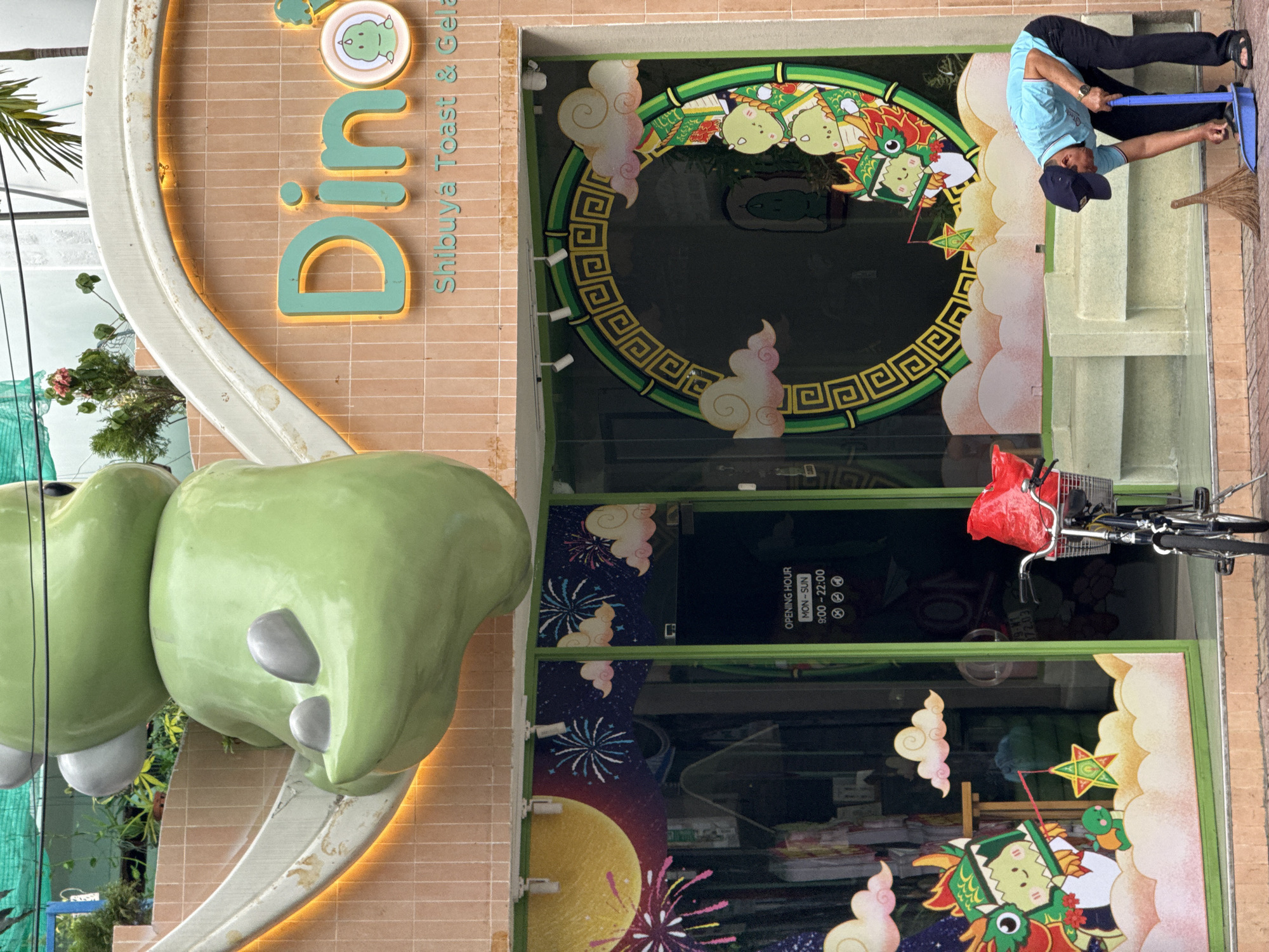 A man is sweeping the sidewalk in front of a storefront in Nha Trang, Vietnam. The storefront has a large, cartoonish green dragon statue above the entrance and a "Dino" sign with "Shibuya Toast & Gelato" written below. The windows are decorated with cartoon dragons, clouds, and fireworks. A bicycle with a red bag in its basket is parked near the entrance. The man is wearing a blue shirt, black pants, a dark baseball cap, and sandals. He is holding a broom and dustpan, actively sweeping. The environment appears to be a street scene with a building facade.