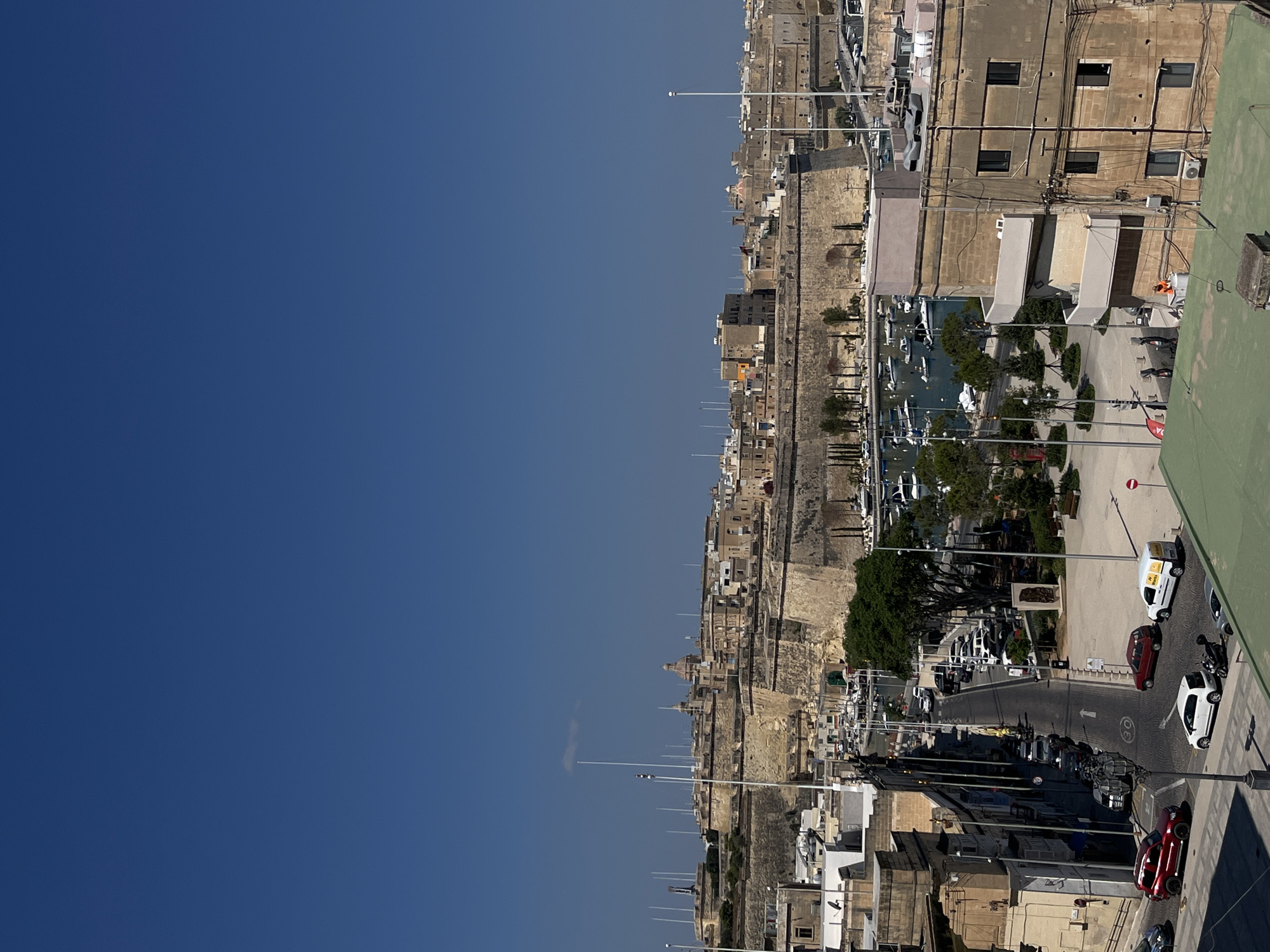 This image shows a view of a harbor and historic fortifications under a clear blue sky. The foreground features a street with several parked cars, including a red SUV, a white hatchback, and a white van. A motorcycle is also visible. In the mid-ground, a harbor is filled with moored boats of various sizes. Beyond the harbor, massive stone fortifications dominate the landscape, with buildings nestled within and behind them. Tall poles, likely for flags or antennas, are scattered across the fortifications and the city skyline. In the background, more buildings and structures extend into the distance. The overall scene depicts a sunny day in a coastal city with significant historical architecture and a busy waterfront.