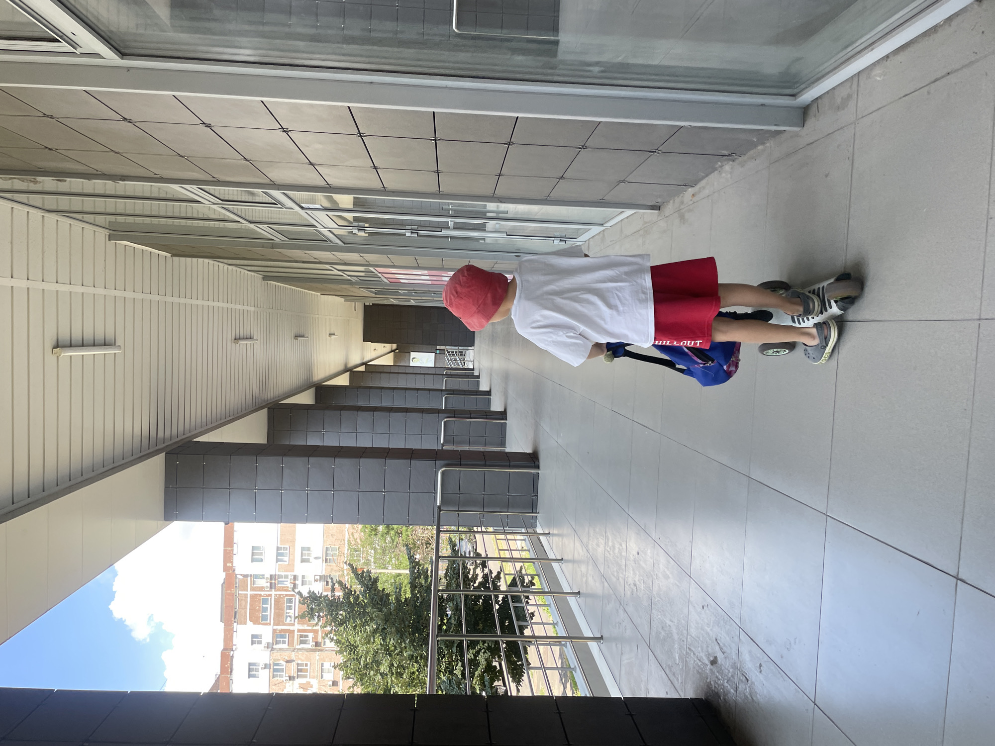 A young child, viewed from behind, rides a scooter down a tiled outdoor walkway. The child wears a red bucket hat, a white t-shirt, and red shorts, and carries a blue bag over their shoulder. The walkway is lined with large, dark grey rectangular panels and reflective windows on one side, and a metal railing with a view of trees and a residential building on the other. The scene appears to be in an urban setting, possibly Novorossiysk, Russia, given the architectural style and vegetation. The sky is partly cloudy and blue.
