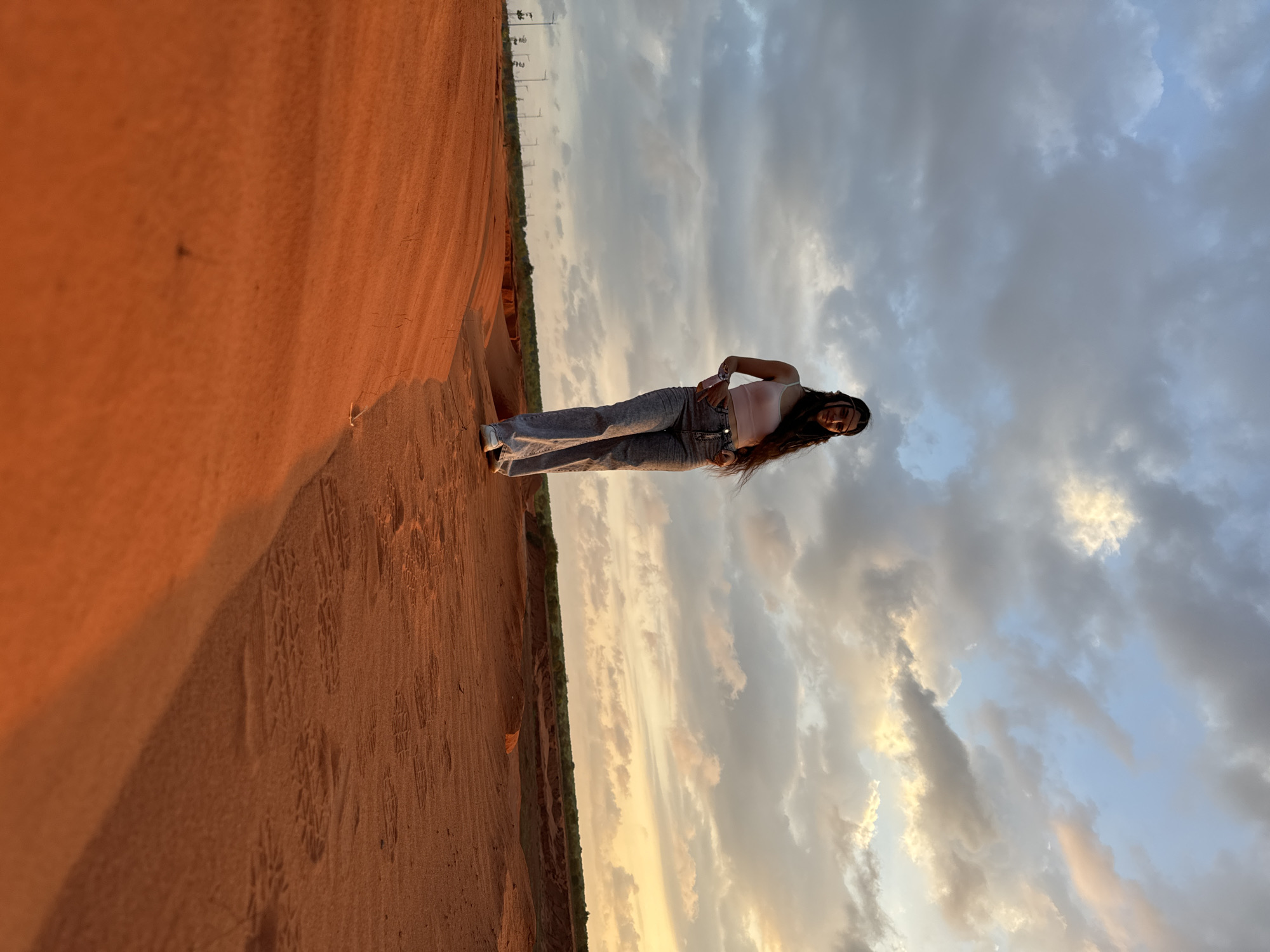 A young woman stands in the Mui Ne sand dunes, Vietnam. The sky is filled with clouds and the sun is starting to set, creating a warm and orange glow. The dunes are an orange-red color, and there are some tracks visible in the sand. The woman is wearing a light pink top and gray jeans, and her hair is long and dark. She is posing with one hand on her hip.
